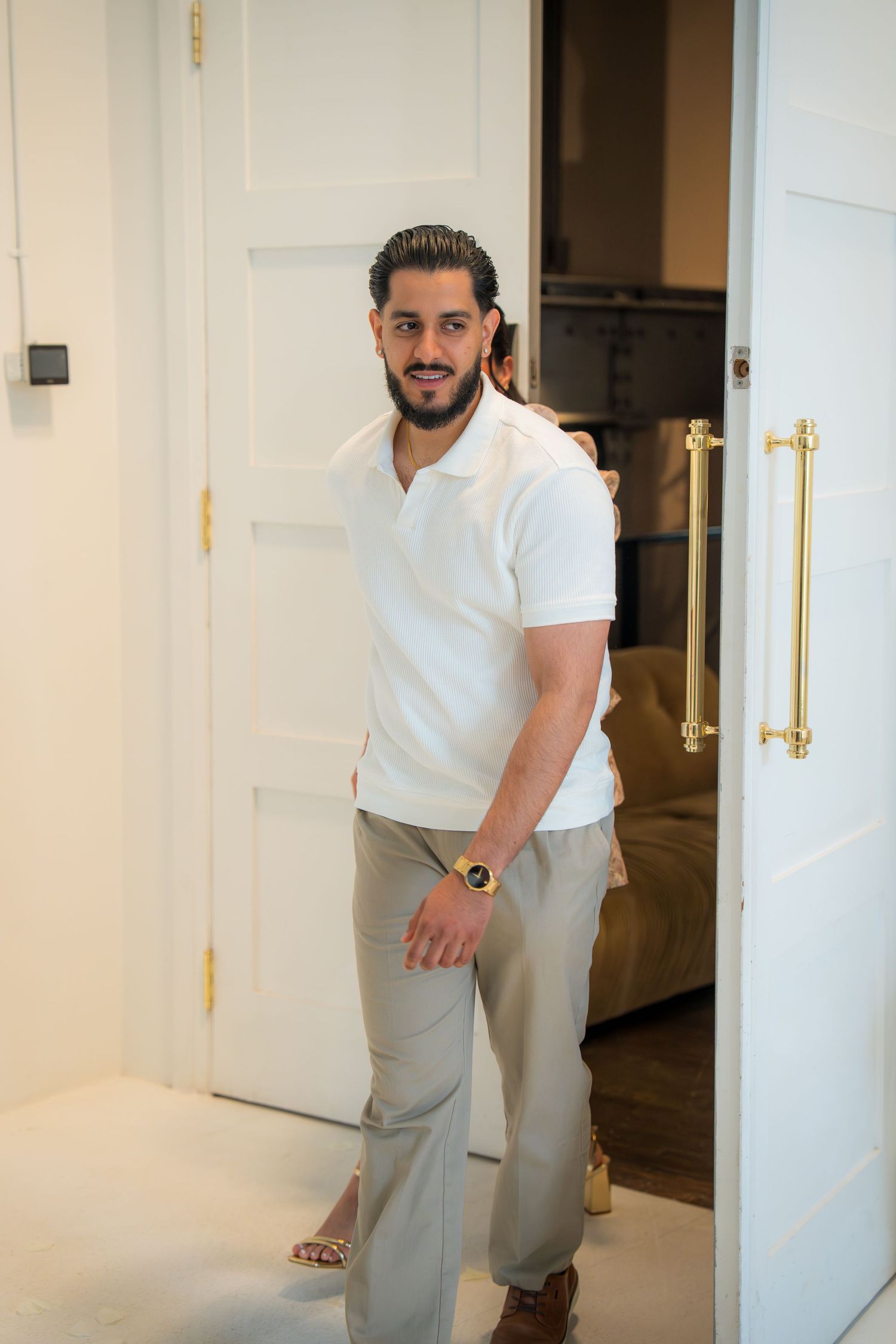 Stylish man in white polo and khaki pants standing in an elegant doorway with gold hardware.