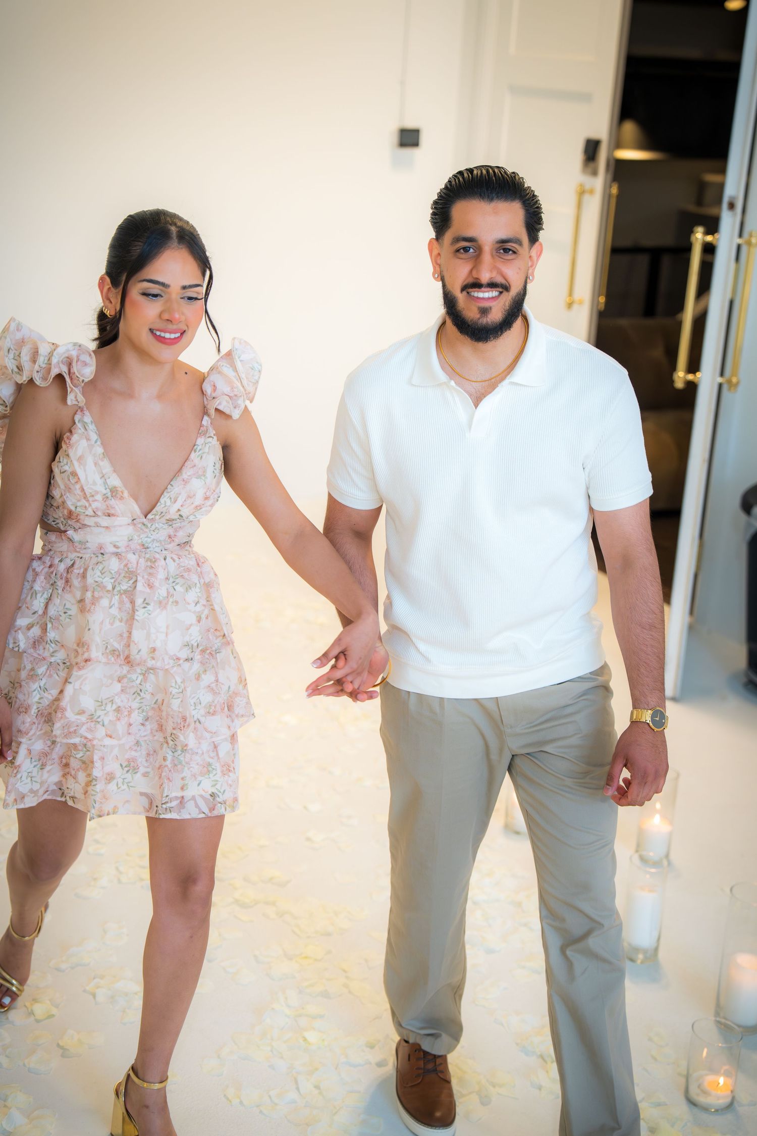 Smiling couple holding hands walking through a romantic white floral proposal setup with candles.