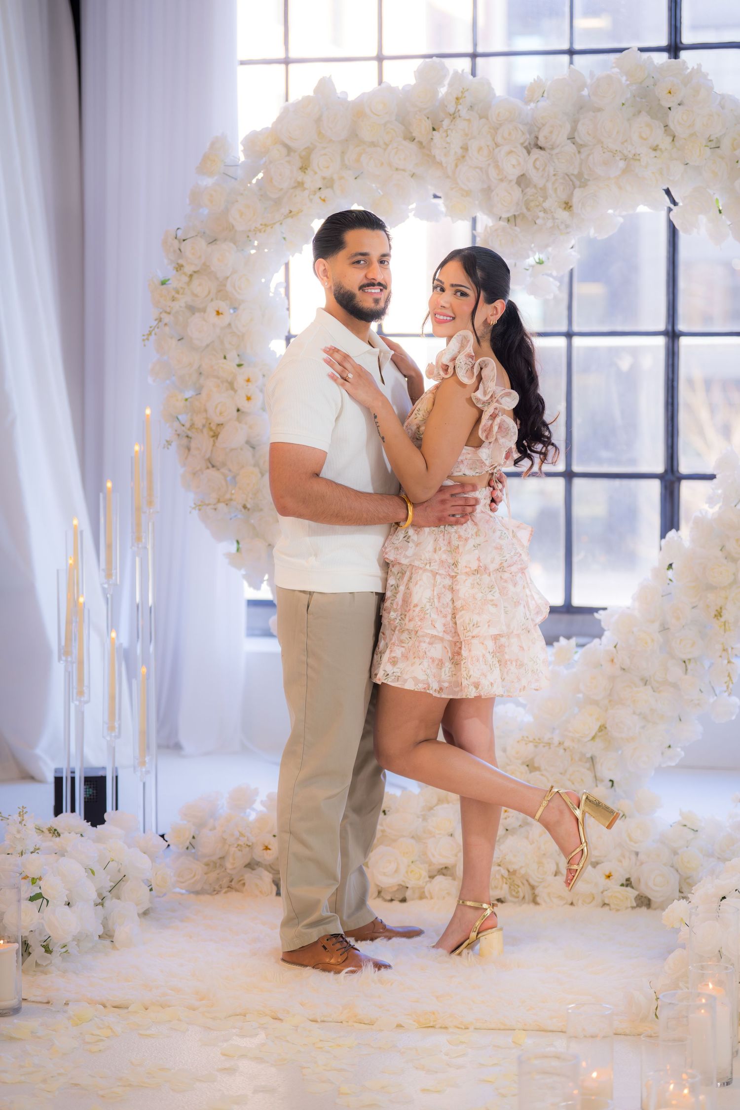 Happy couple posing together in front of stunning white heart-shaped flower arch with candles.