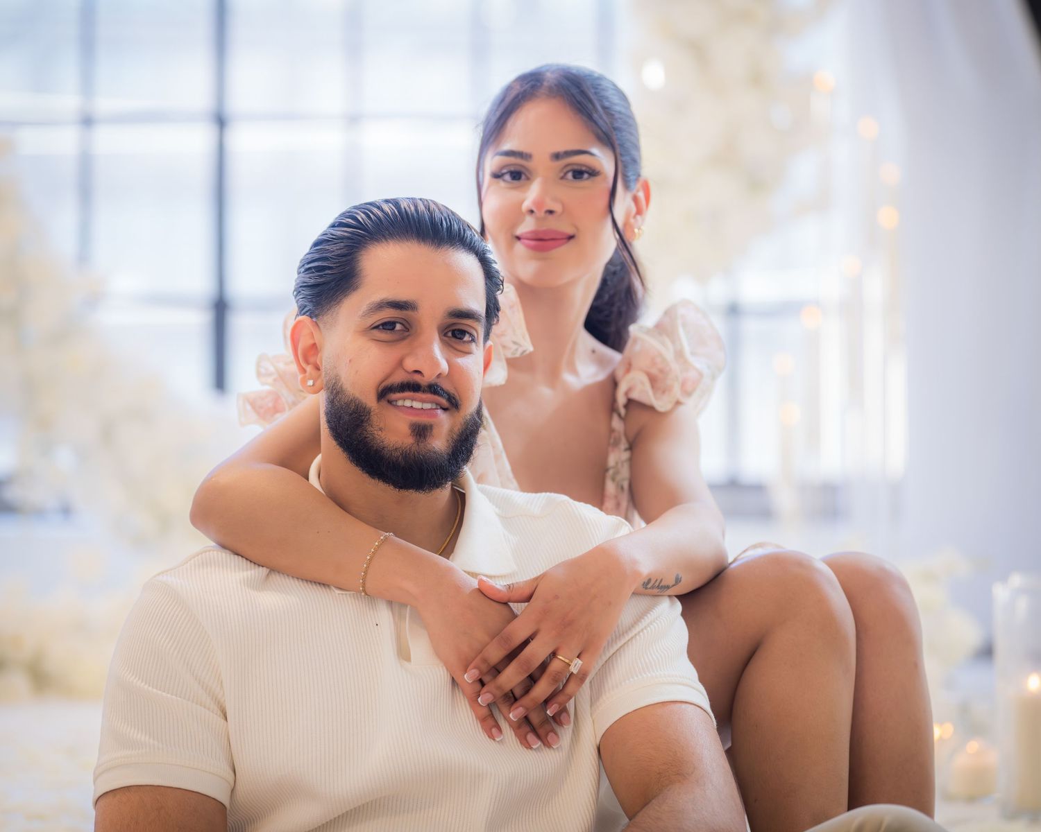 Engaged couple posing together surrounded by white floral decorations and candles.