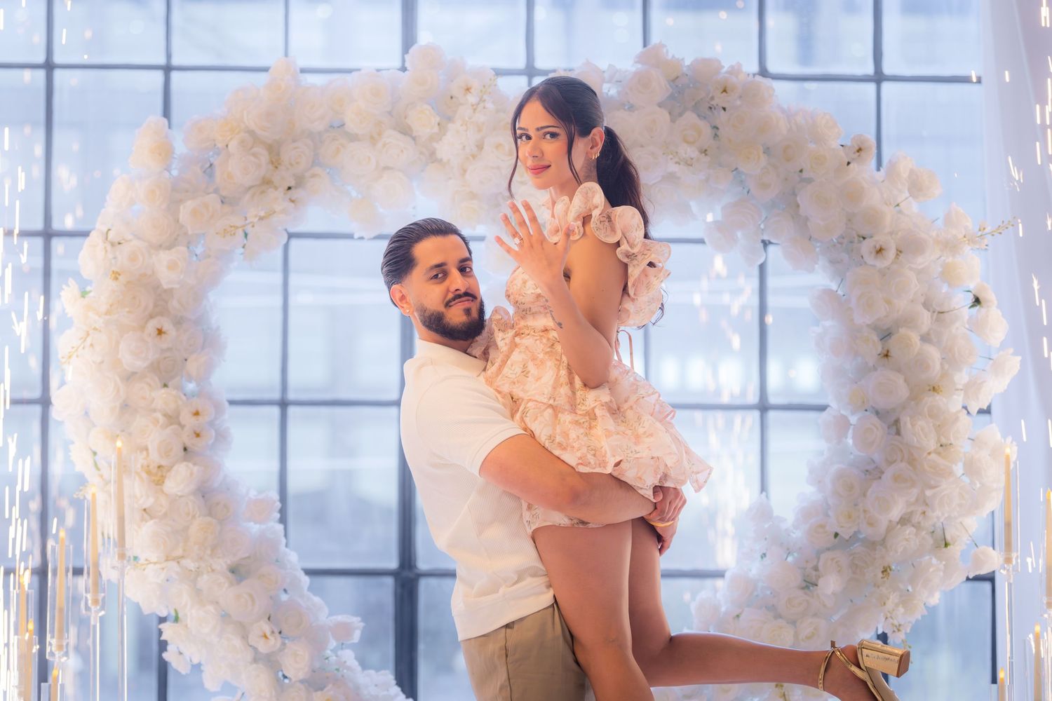 Man lifting woman joyfully in front of a large white circular floral arch backdrop.