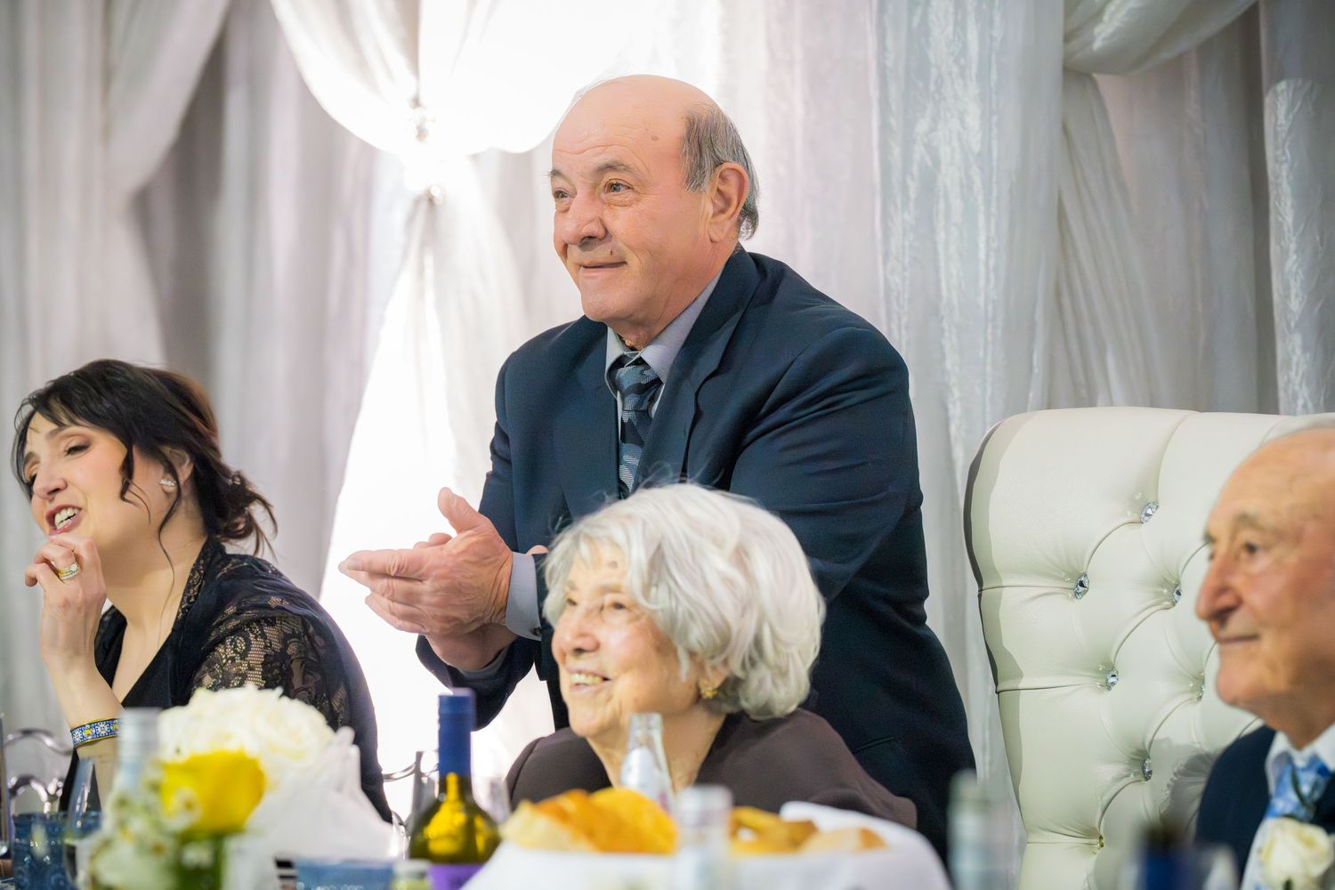 Older gentleman in navy suit speaking animatedly at a formal banquet table with yellow and white floral centerpieces.