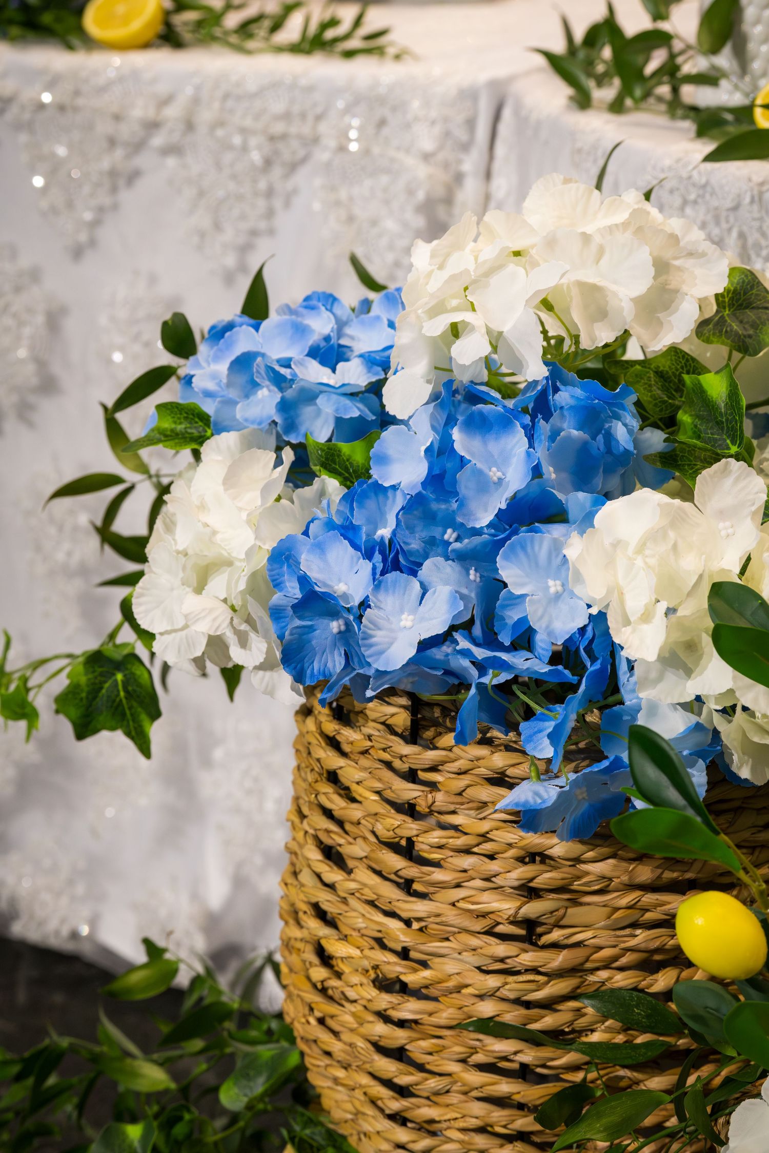 Wicker basket arrangement of blue and white hydrangeas with green leaves and fresh lemons for elegant event decor.