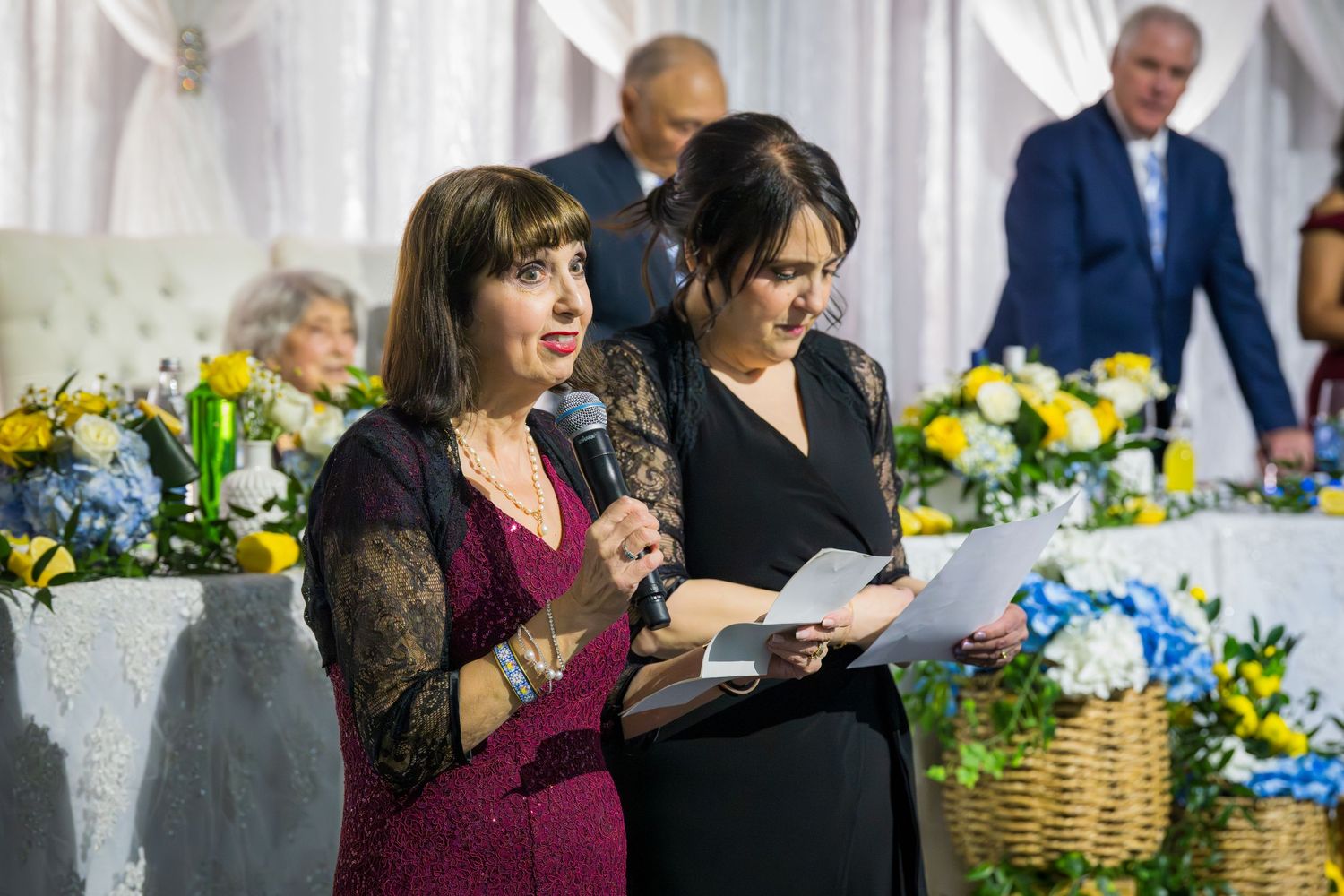 Two women in formal attire speak into a microphone at a celebration surrounded by yellow and blue floral arrangements.