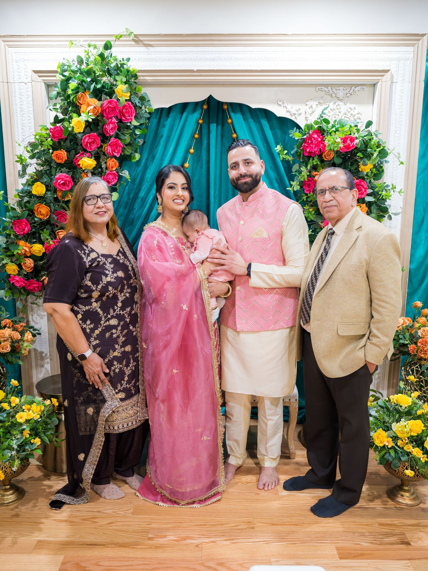 Family portrait with parents holding baby in pink traditional attire before vibrant floral Lohri backdrop.