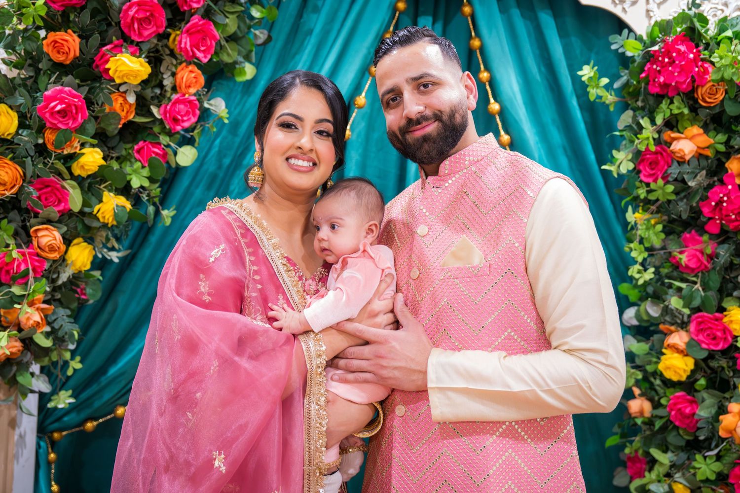 Smiling South Asian couple in pink traditional dress holds infant at vibrant flower-adorned teal drape.