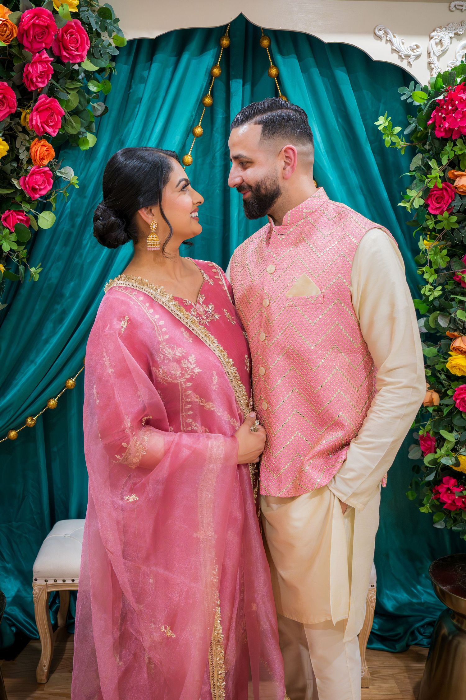 Couple in coordinating pink and gold South Asian traditional attire gazing lovingly at each other at a festive event.