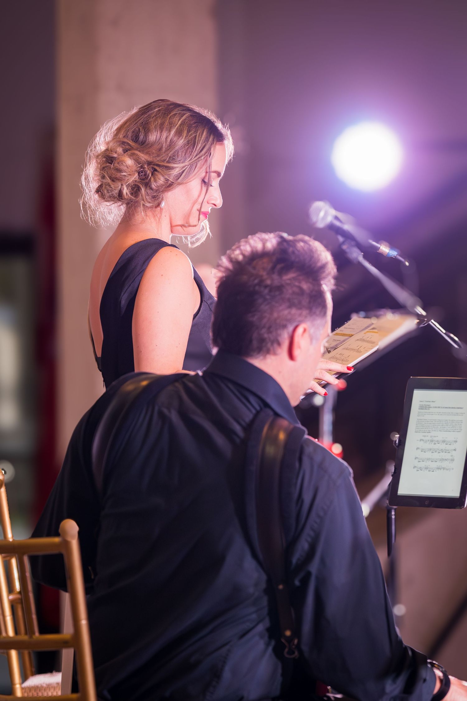 Female vocalist in black dress reviews sheet music with male guitarist on stage under warm lights.