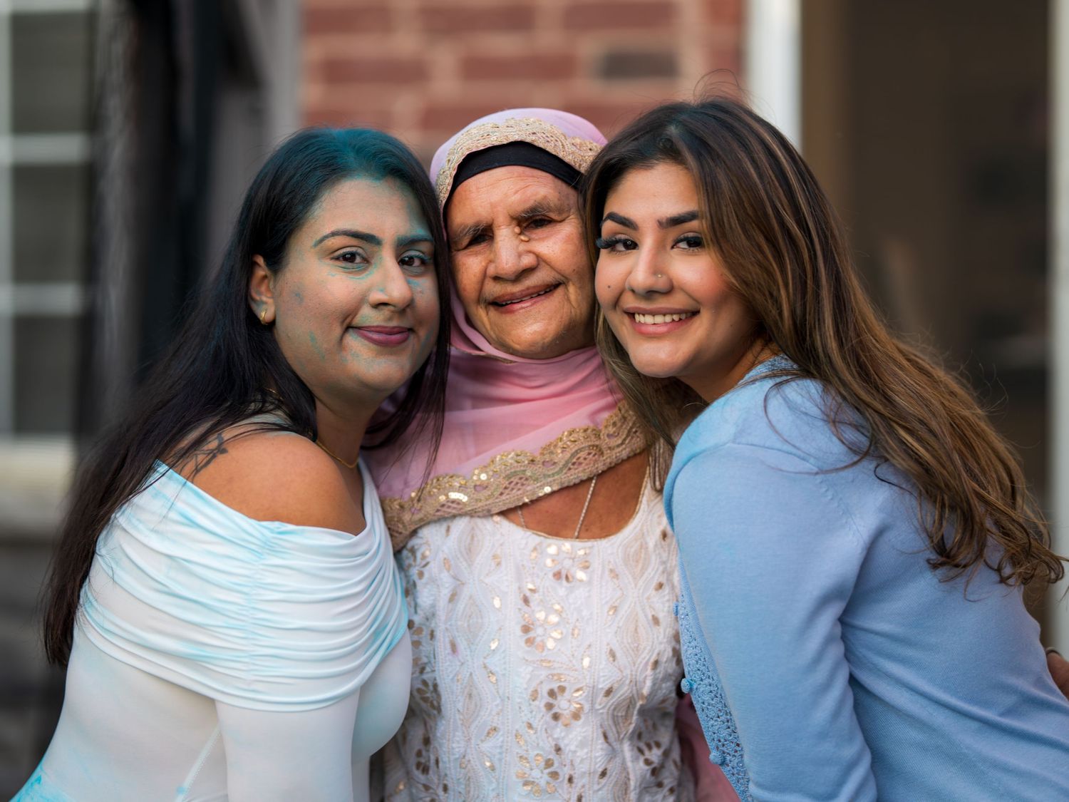 Three women smile together for a photo, with the center woman wearing a pink hijab and white outfit.