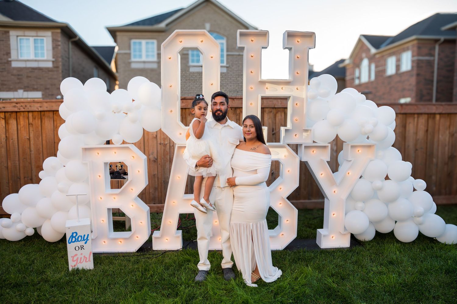 A family of three poses in white outfits before an OH BABY balloon and marquee display.