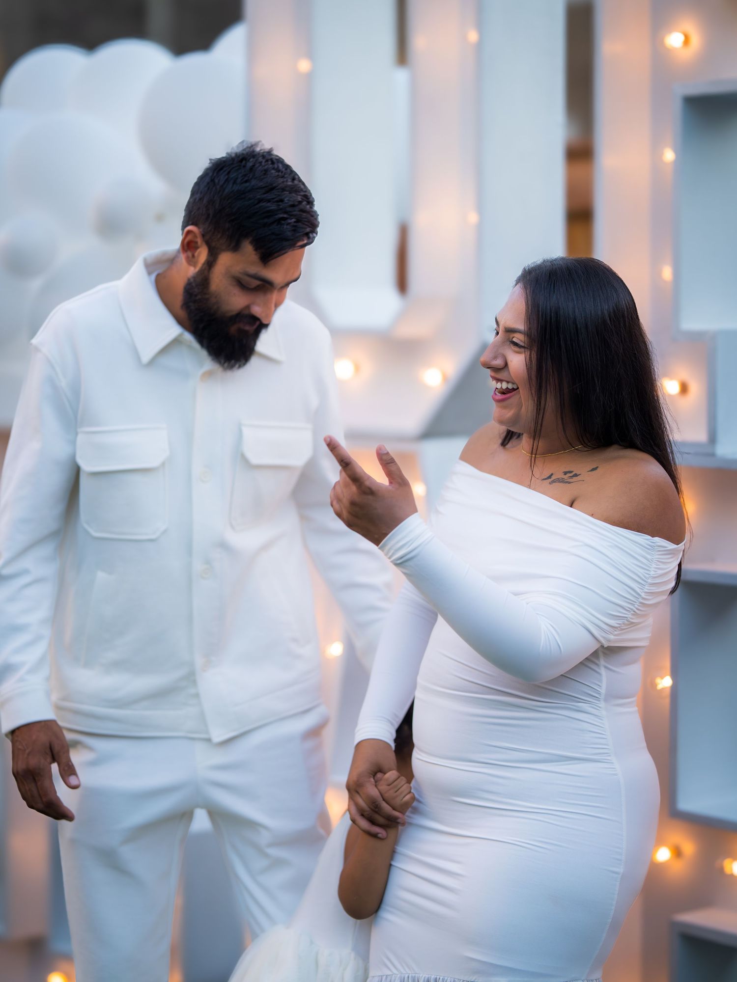 Expecting couple in matching white outfits smiling at gender reveal party with lights.
