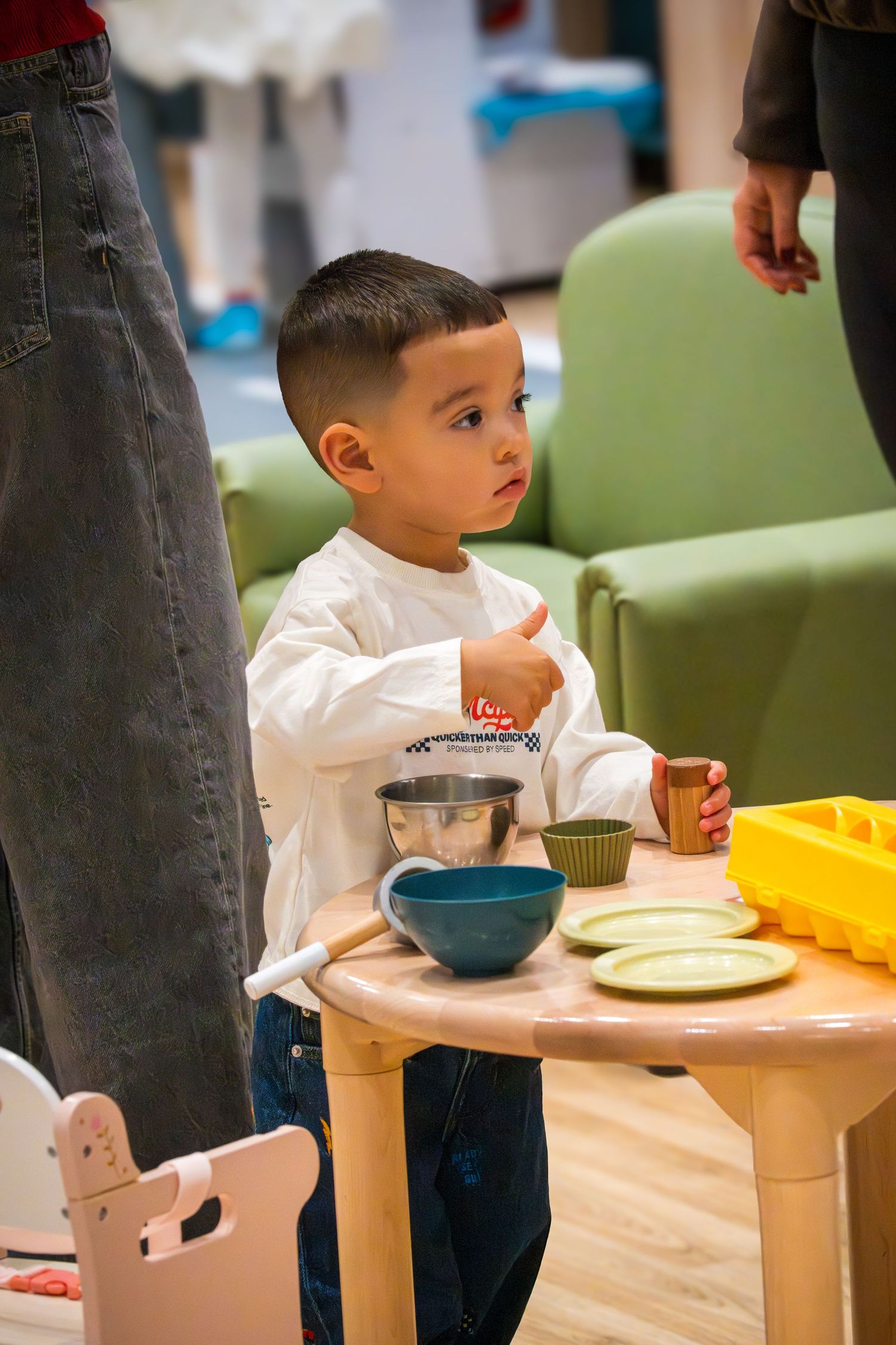 Young boy playing with toy kitchen set on small wooden table, holding cup with bowls and plates nearby.