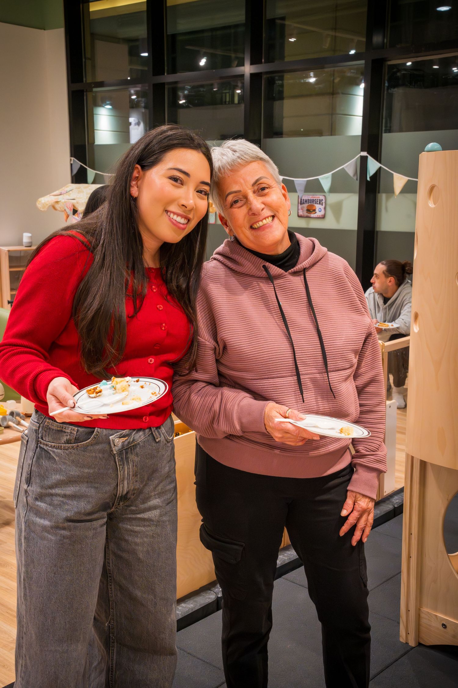 Two women smiling and holding plates of food at an indoor gathering or party event.