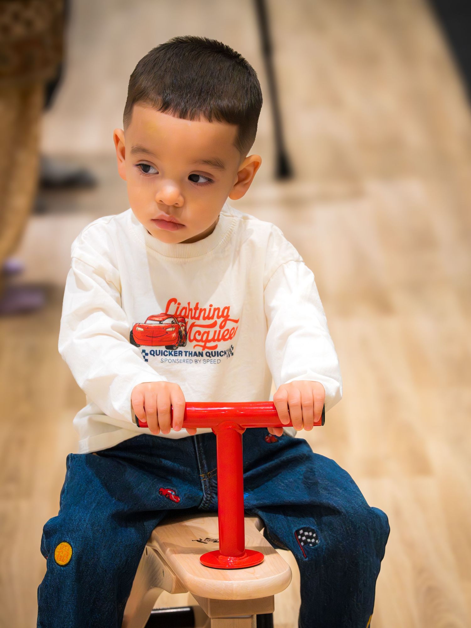 Toddler boy in Lightning McQueen shirt riding a wooden balance bike on hardwood floor.