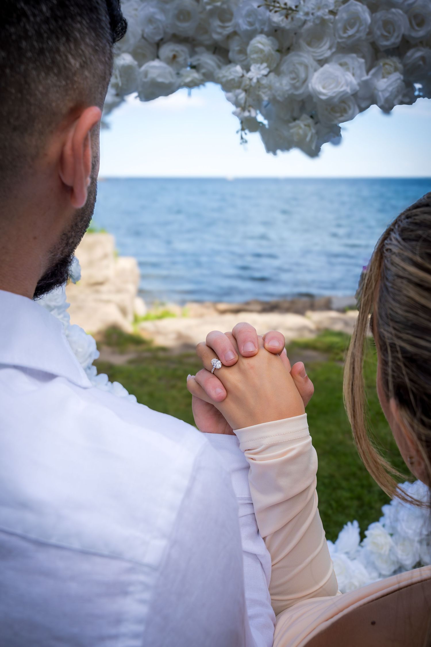 Couple holding hands during a seaside proposal beneath a white floral arch.