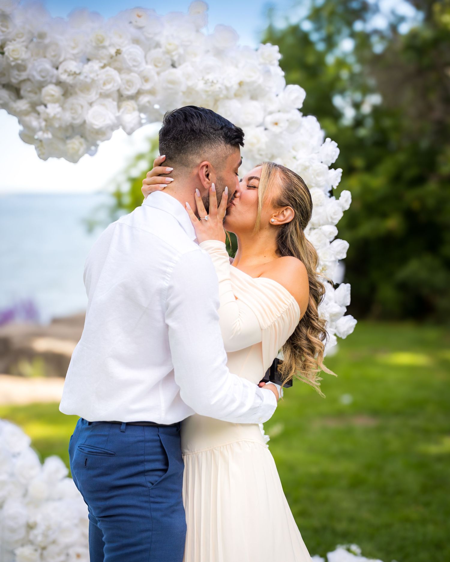 Intimate proposal moment under decorative white flower arch in outdoor garden setting.
