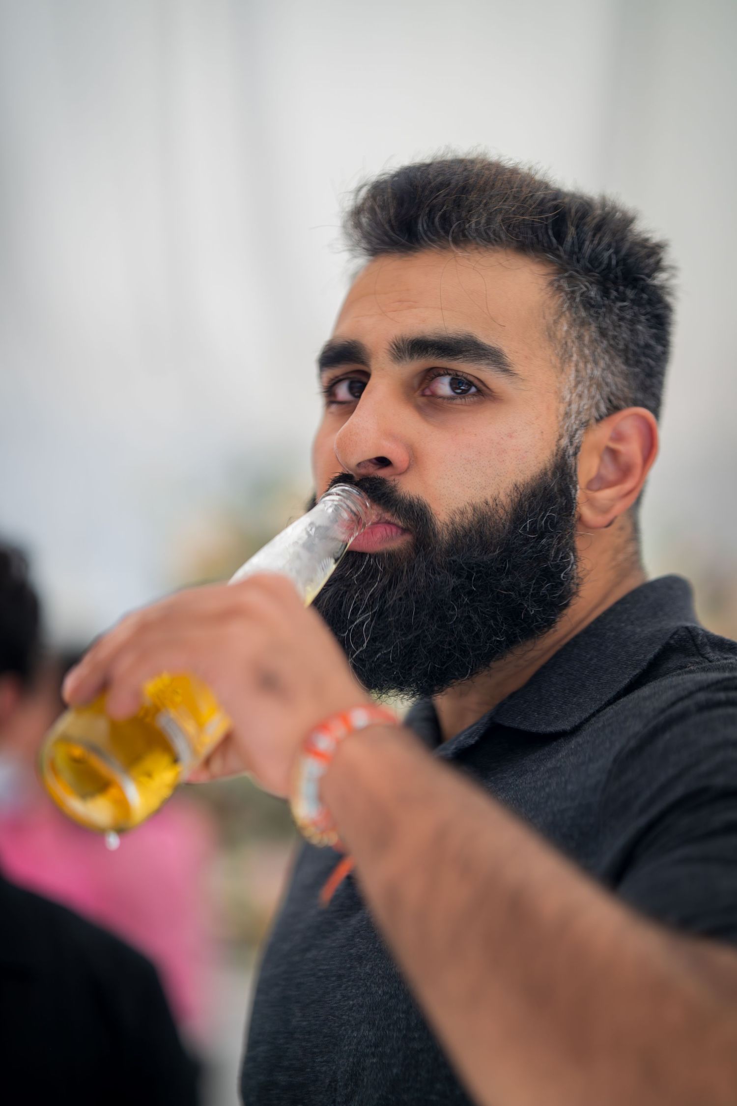 A person with a dark beard drinks from a beer bottle at a social gathering.