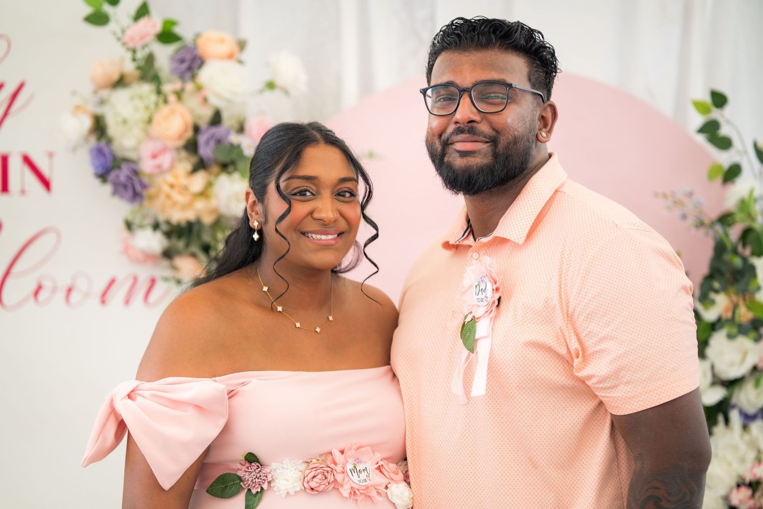 Couple in coordinated pink and peach outfits posing at a baby shower with floral decorations.