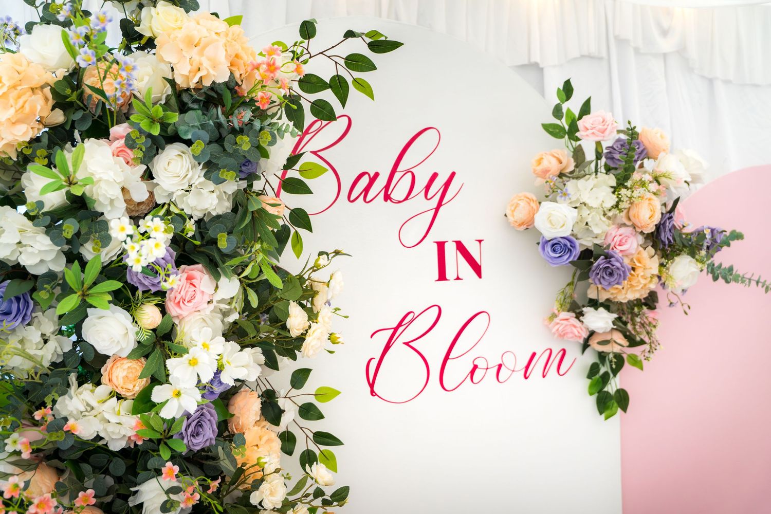 Luxurious baby shower setup with white sofa and floral arch against pink and white backdrop.