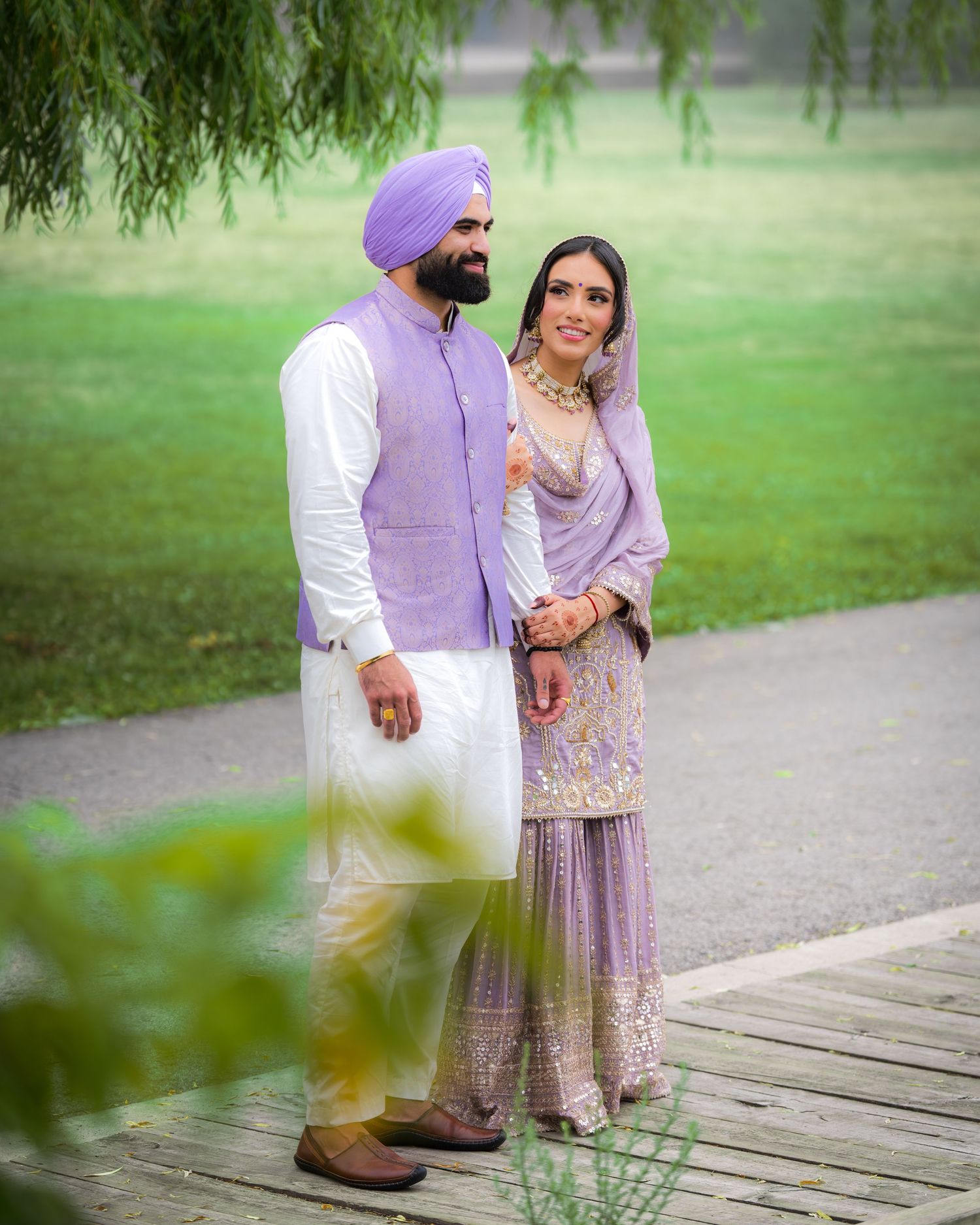 A couple in traditional Punjabi dress posing on a wooden boardwalk in a garden setting.