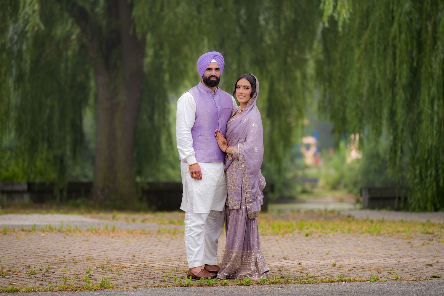 A couple in traditional Punjabi dress stands together on a path beneath drooping willow branches.