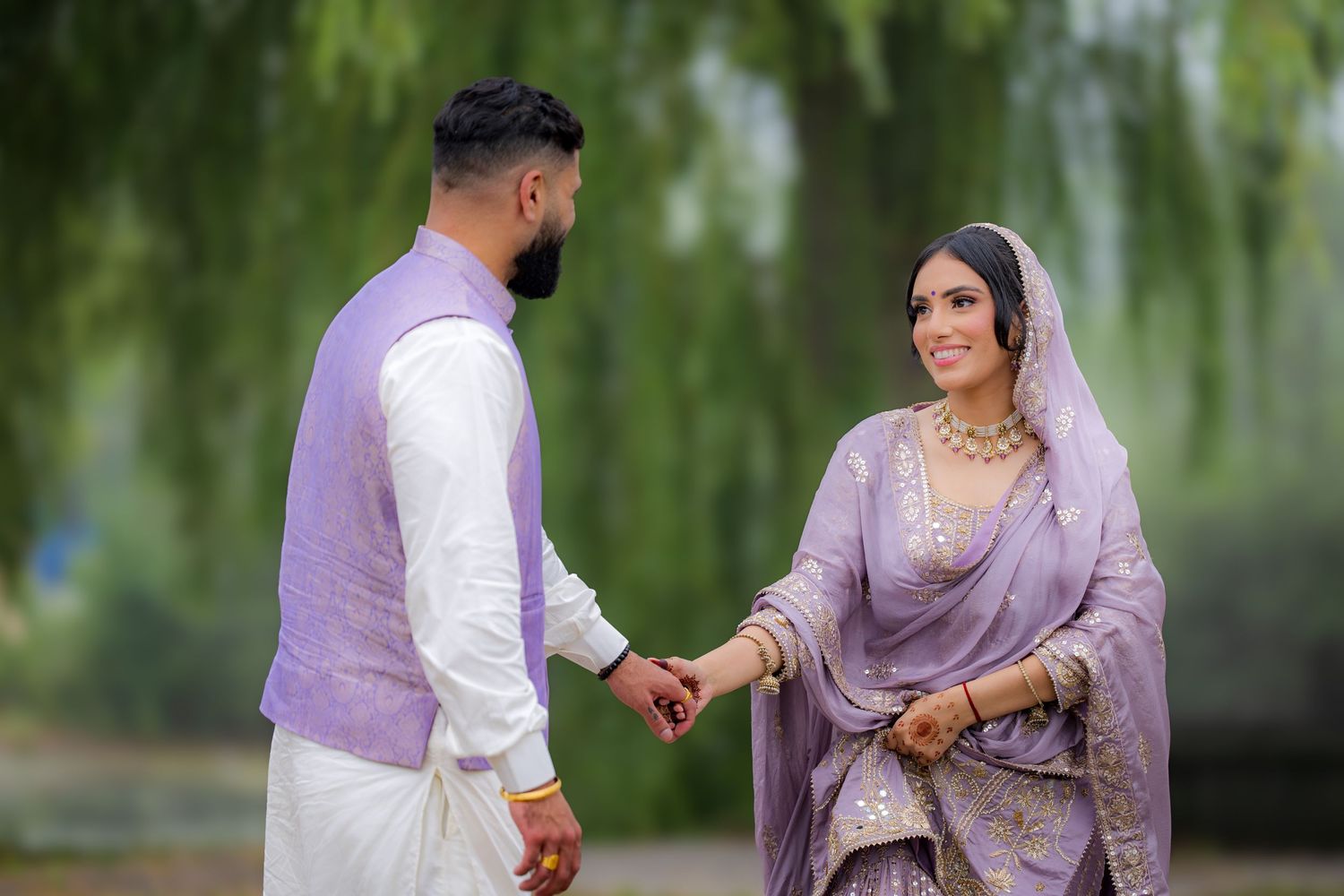 A couple in traditional South Asian wedding attire hold hands against a backdrop of willow trees.