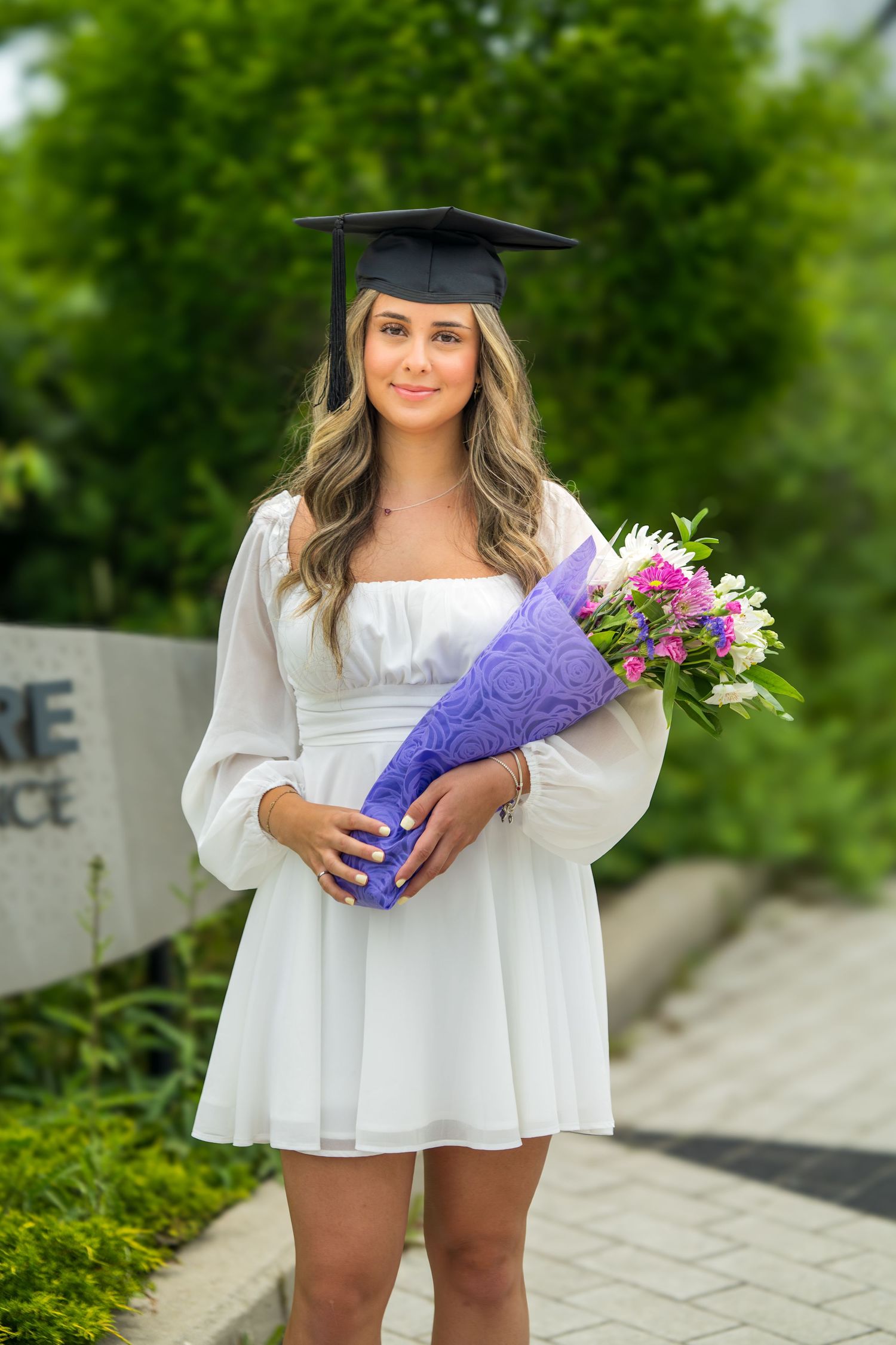 Graduate in white dress and cap holds purple-wrapped flowers on campus pathway.