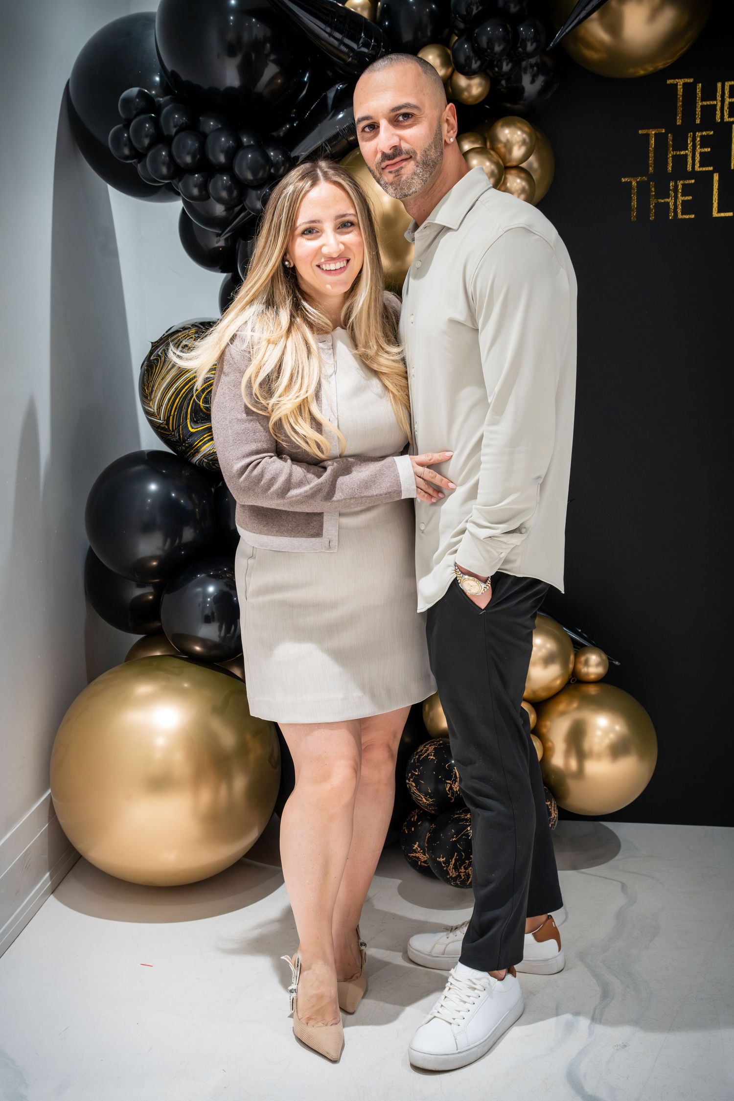 Couple poses together at party against black wall with gold balloon decorations.
