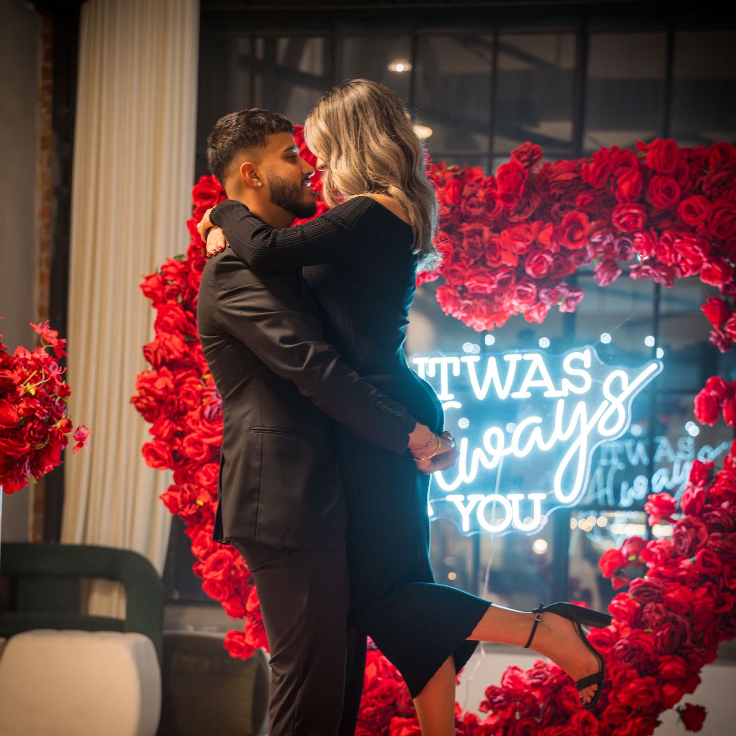 Romantic proposal moment captured against backdrop of red roses and blue neon sign.