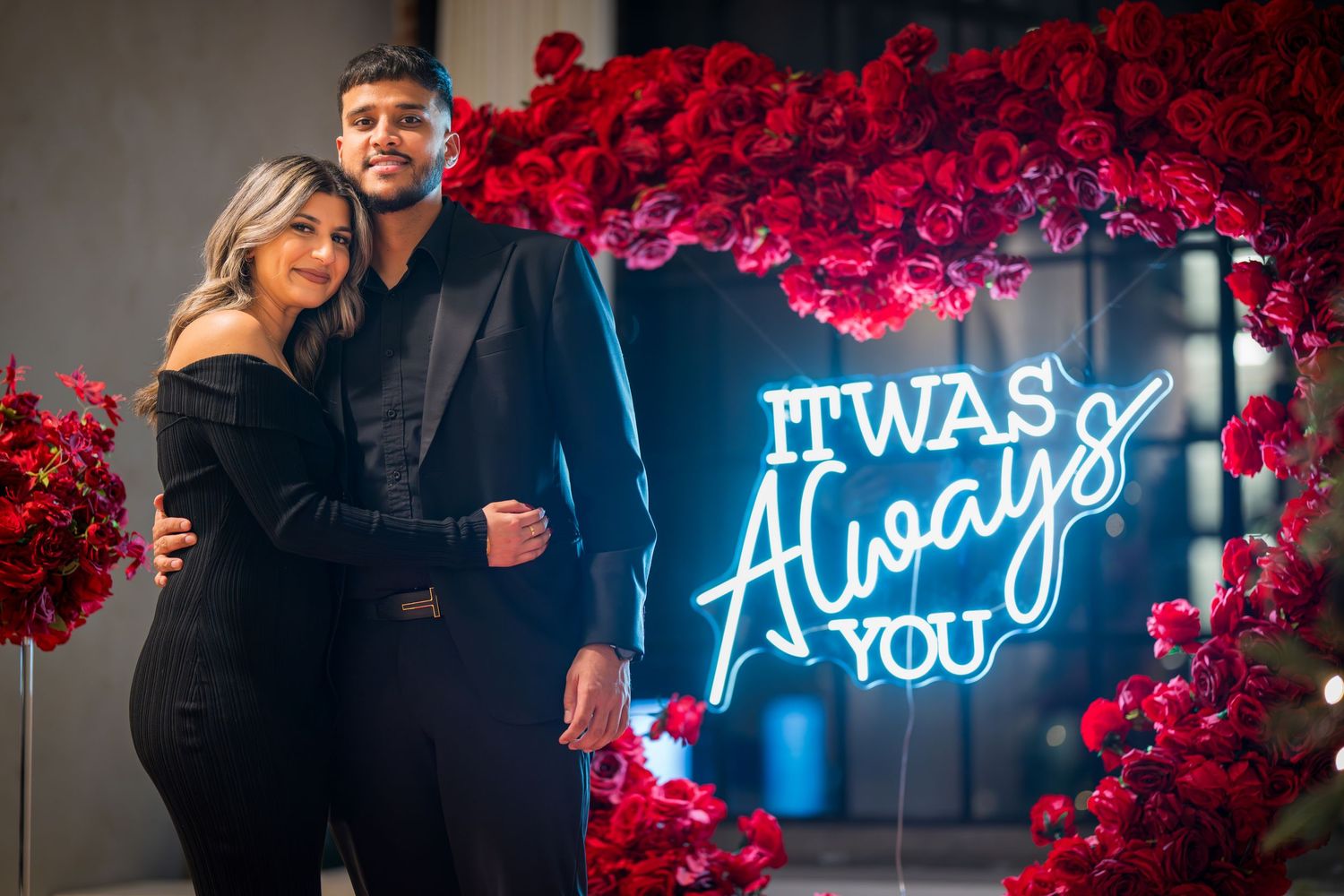 Couple poses elegantly by neon 'It Was Always You' sign surrounded by red roses.