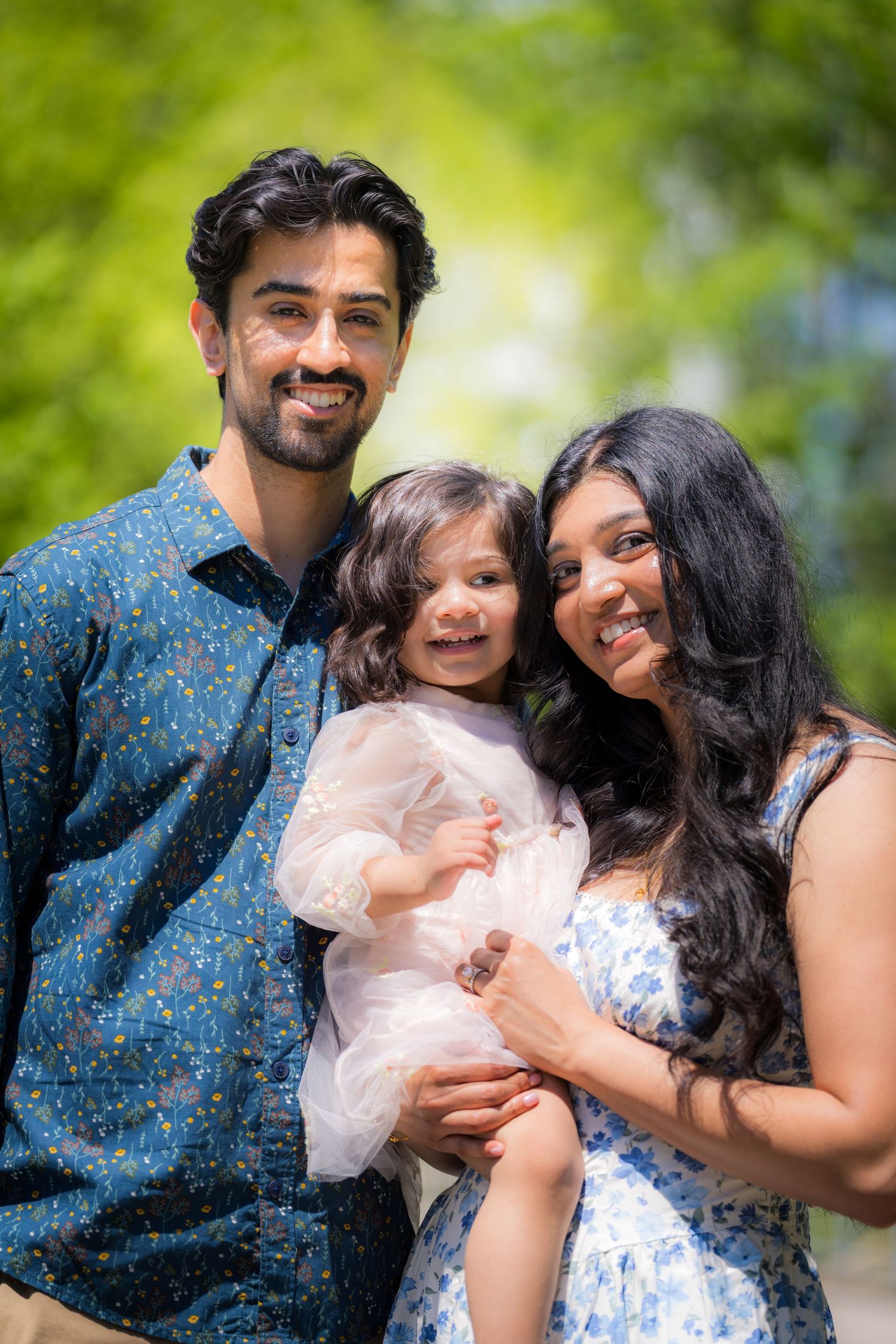 Smiling family of three with toddler girl in white dress pose outdoors in green park.