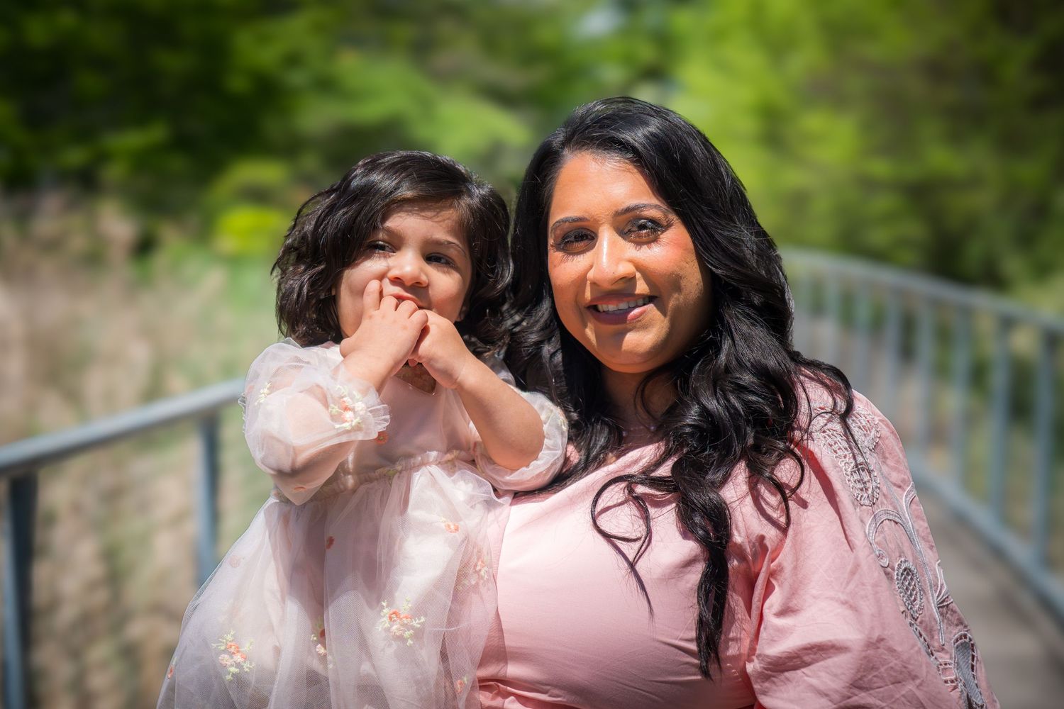 Woman in pink dress poses with young girl in pink tulle outfit on a bridge surrounded by greenery.