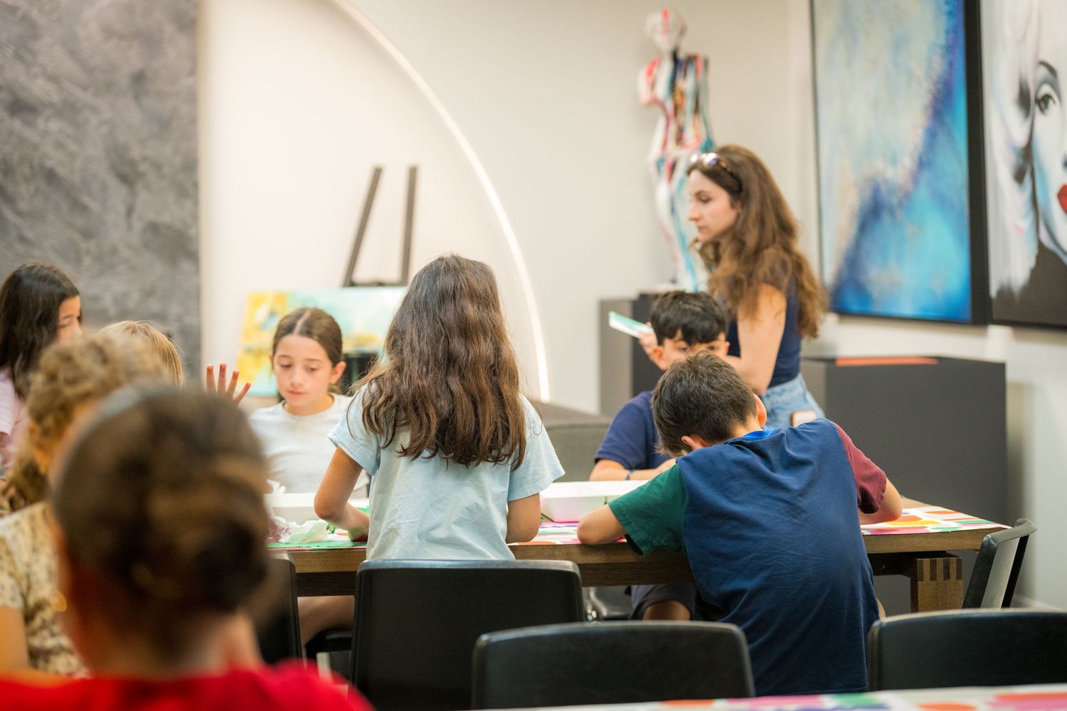 Children sitting at desks in a classroom while a teacher stands and assists students during a lesson.