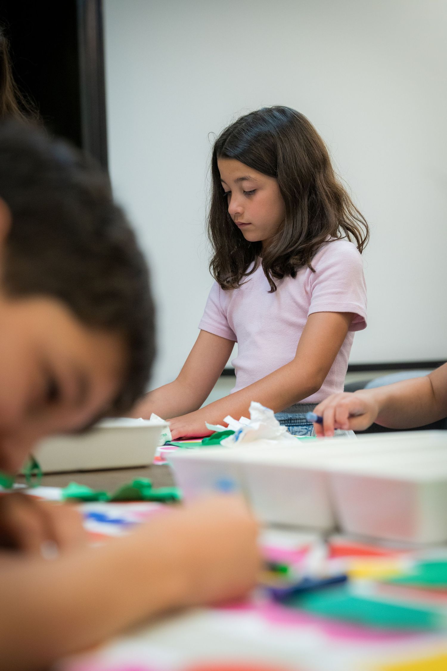 Young girl in pink shirt concentrating on a colorful paper craft activity at a classroom table.