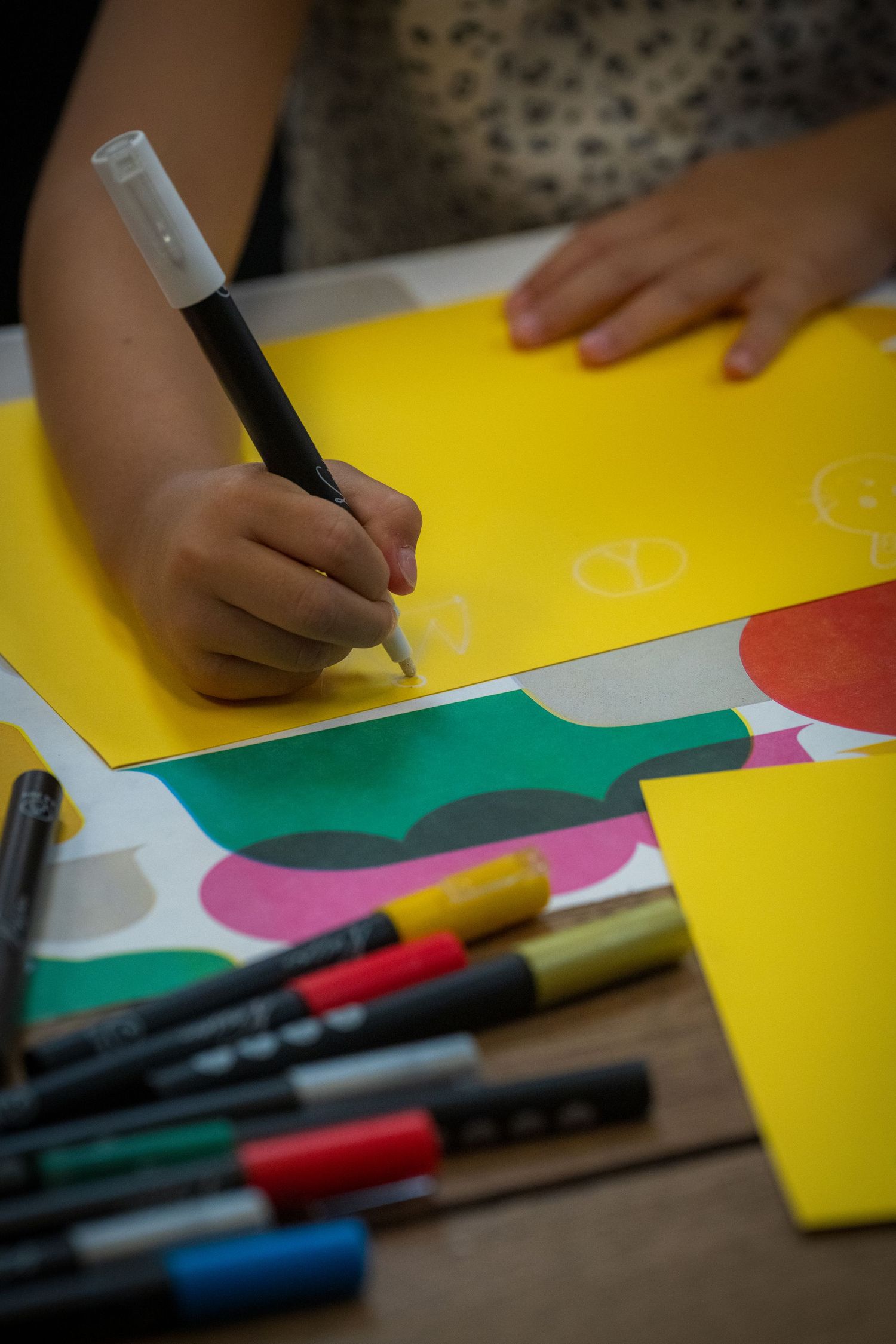 Child's hand writing on yellow paper with black marker surrounded by colorful markers and paper.