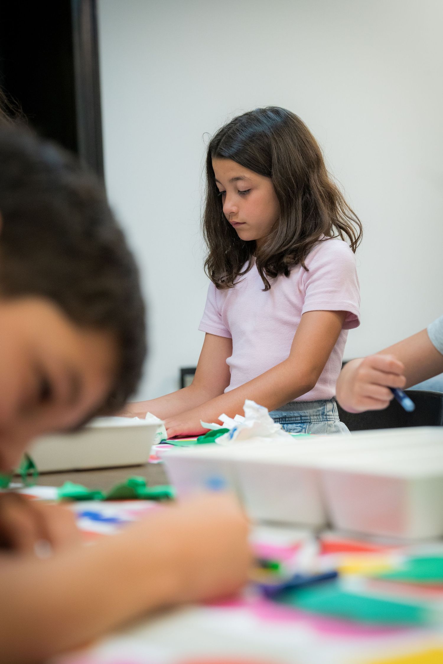 Young girl in a pink shirt focuses on a colorful paper craft activity at a table in an art classroom.