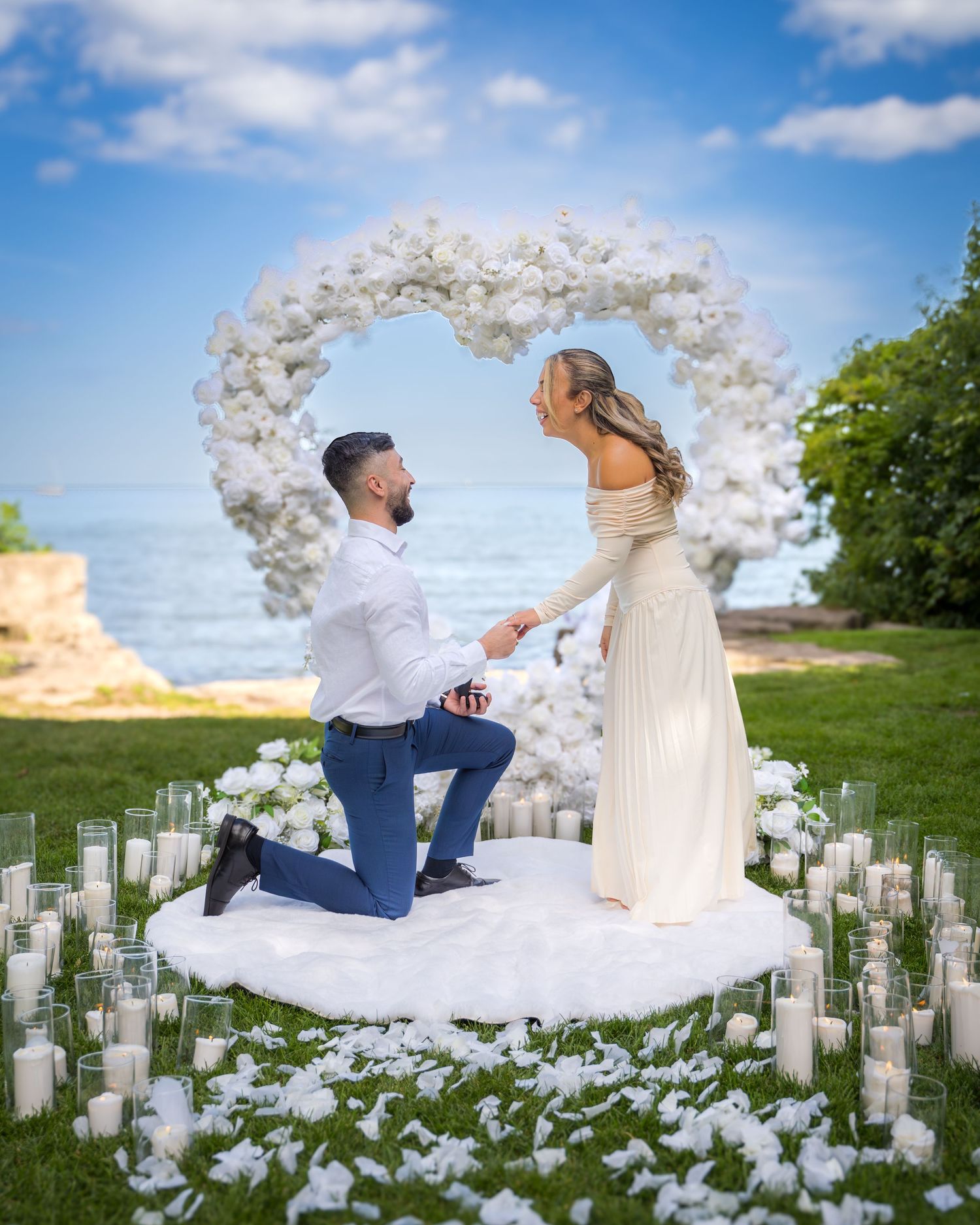 Romantic proposal setup with white balloon arch and candles arranged on grass by the ocean.