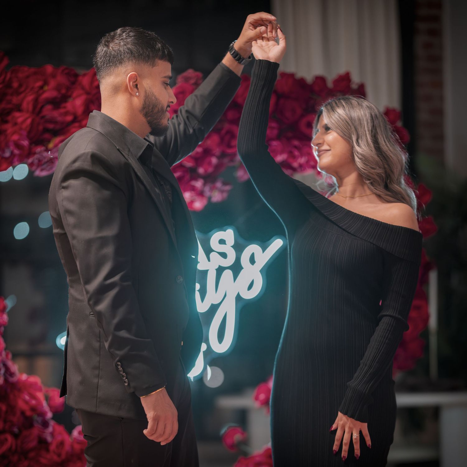Couple dancing in elegant black attire against romantic rose backdrop.