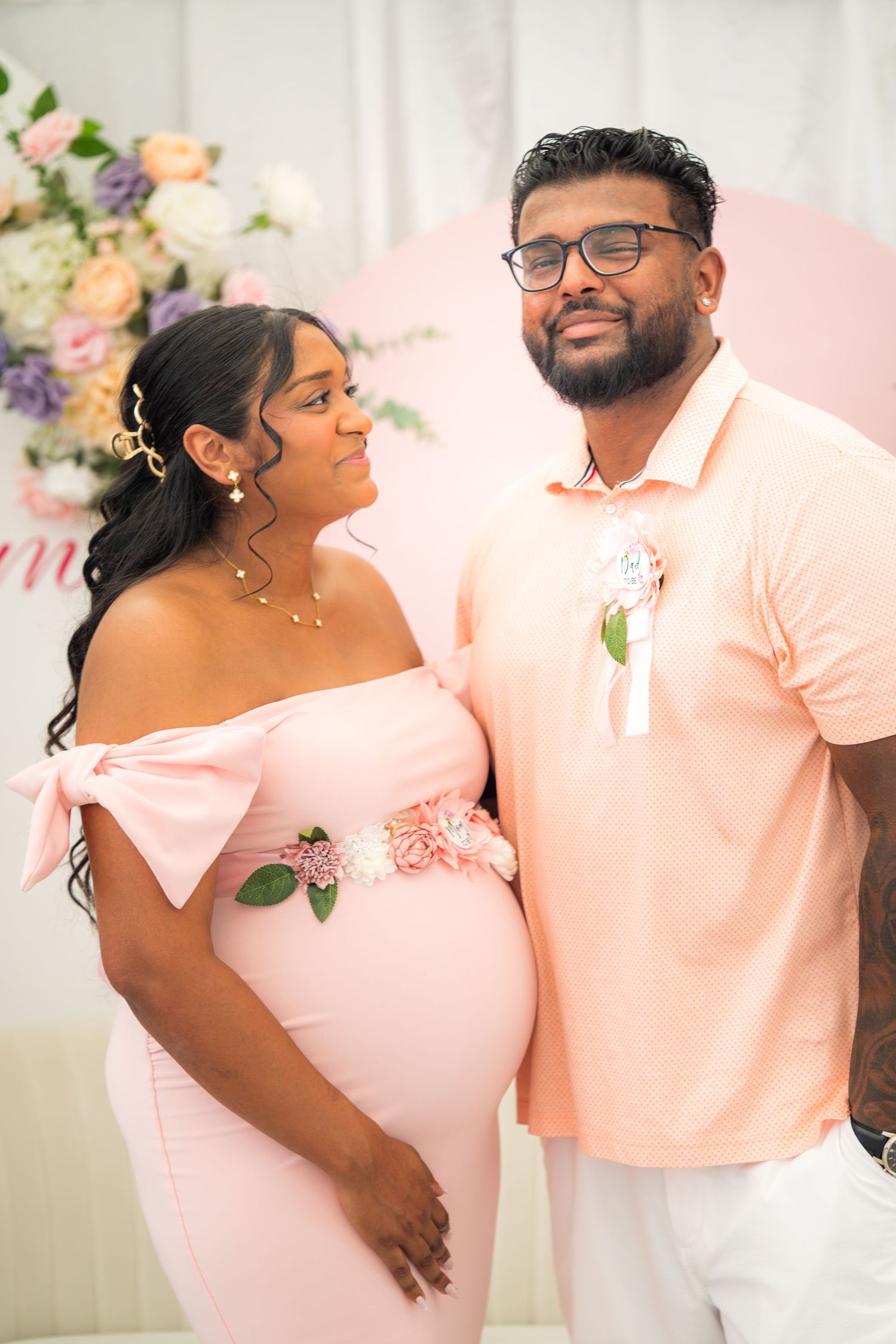 Couple smiles at baby shower celebration with pink floral backdrop and coordinated outfits.