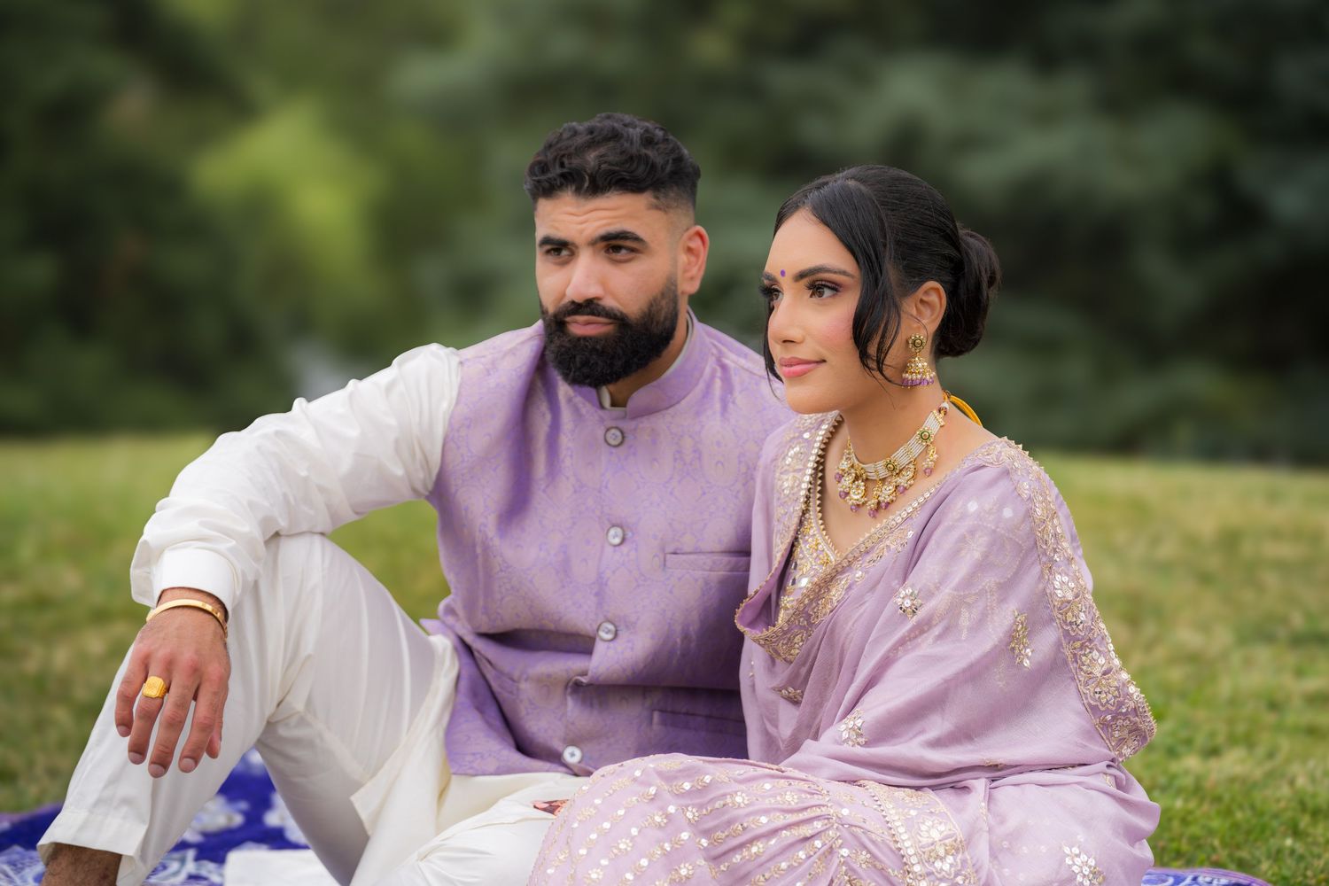 Couple in coordinated lavender and white traditional outfits pose together in an outdoor garden setting.
