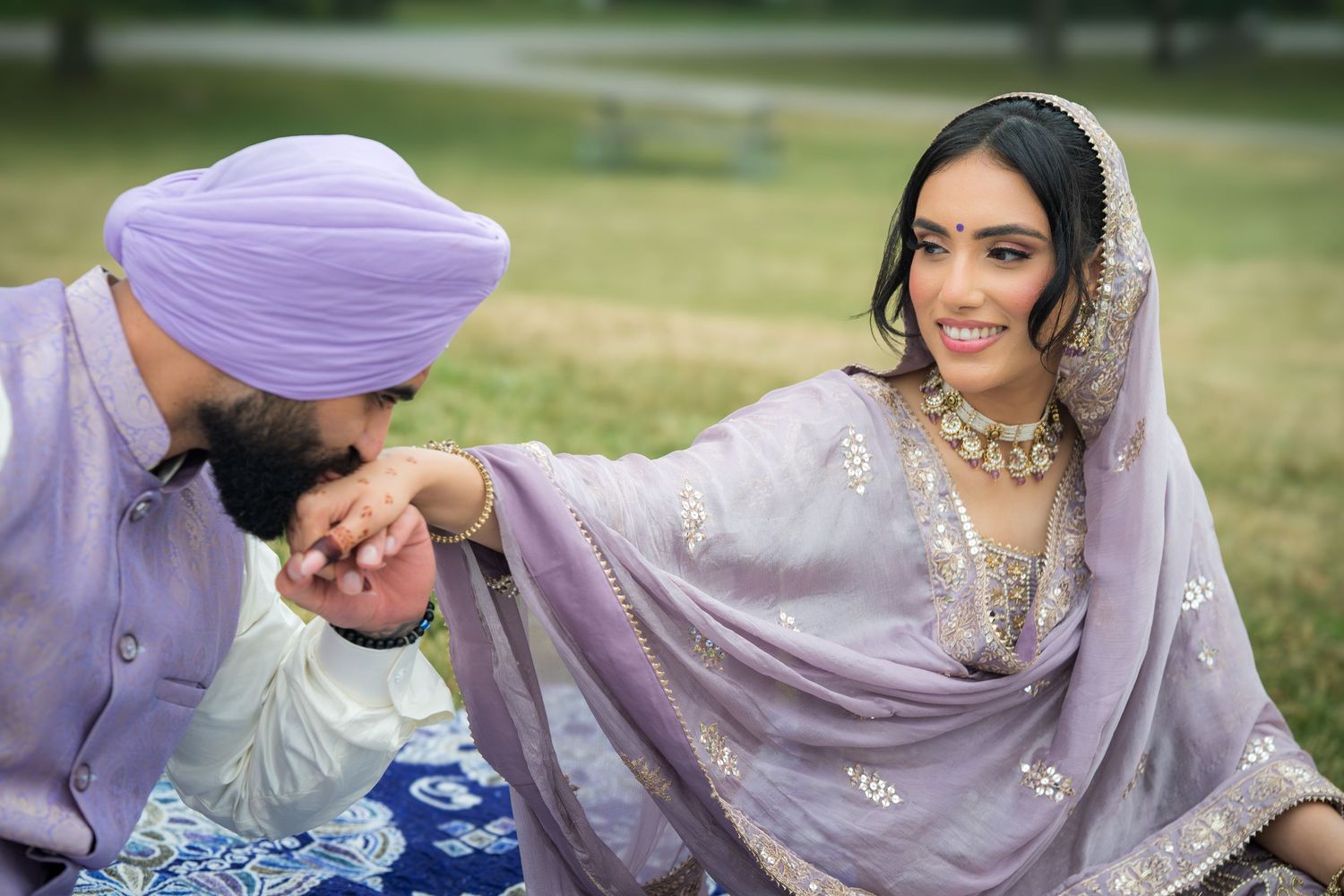 A couple in traditional Punjabi wedding attire poses together outdoors in lavender and white outfits.