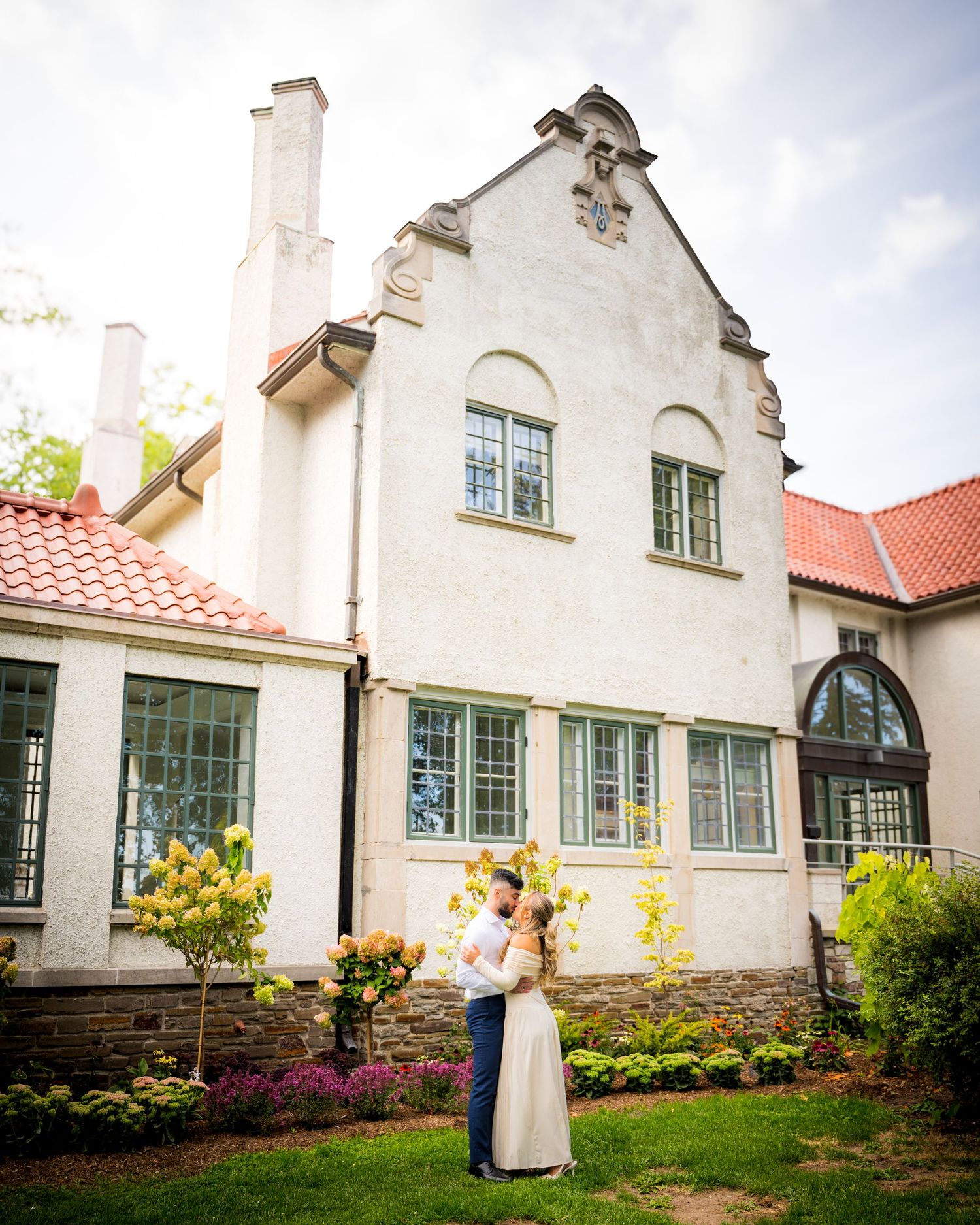 Historic white stucco cottage with Dutch Colonial gables and green windows features a wraparound garden and red tile roof.