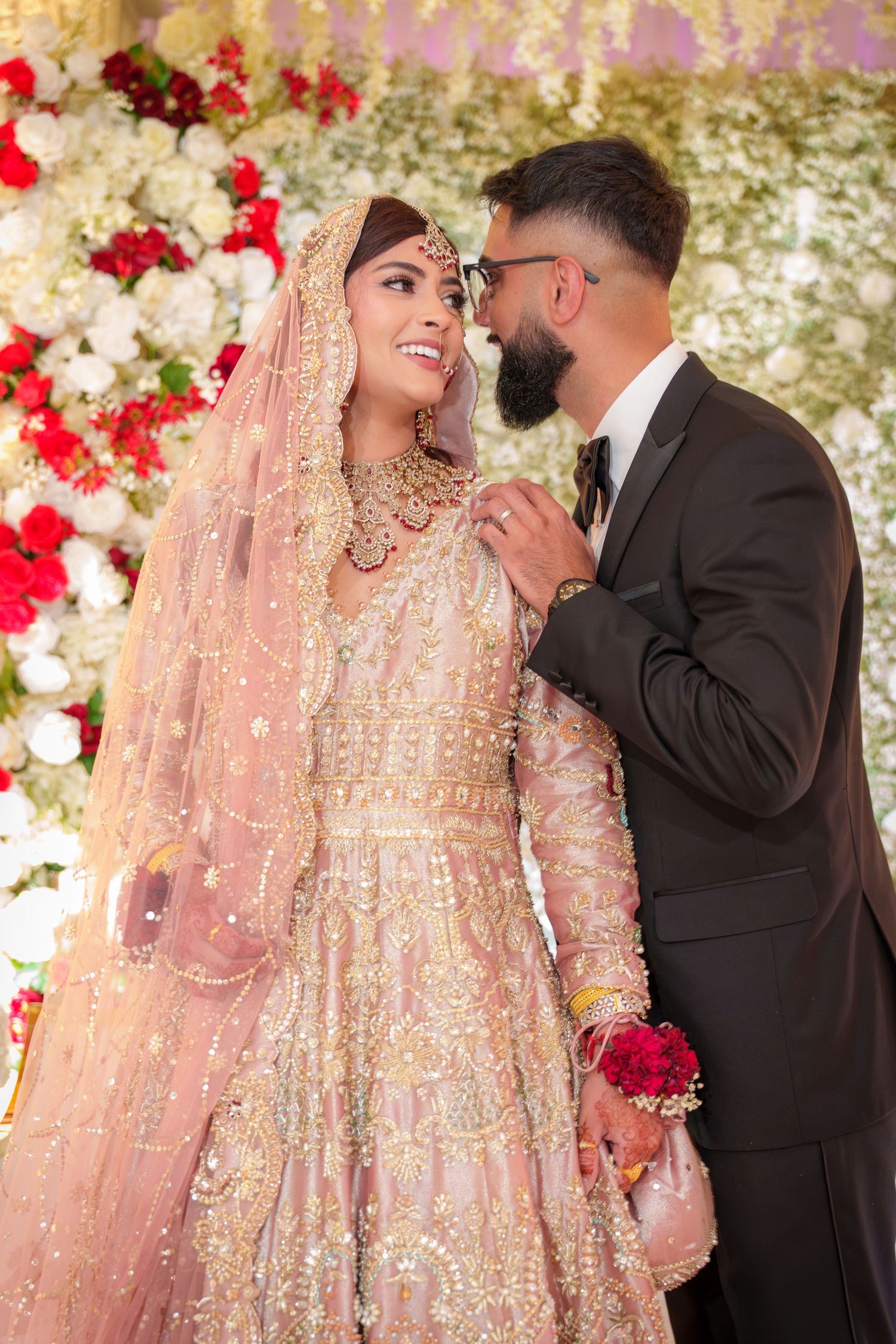 Couple in traditional wedding attire share intimate moment against floral backdrop with red and white roses.