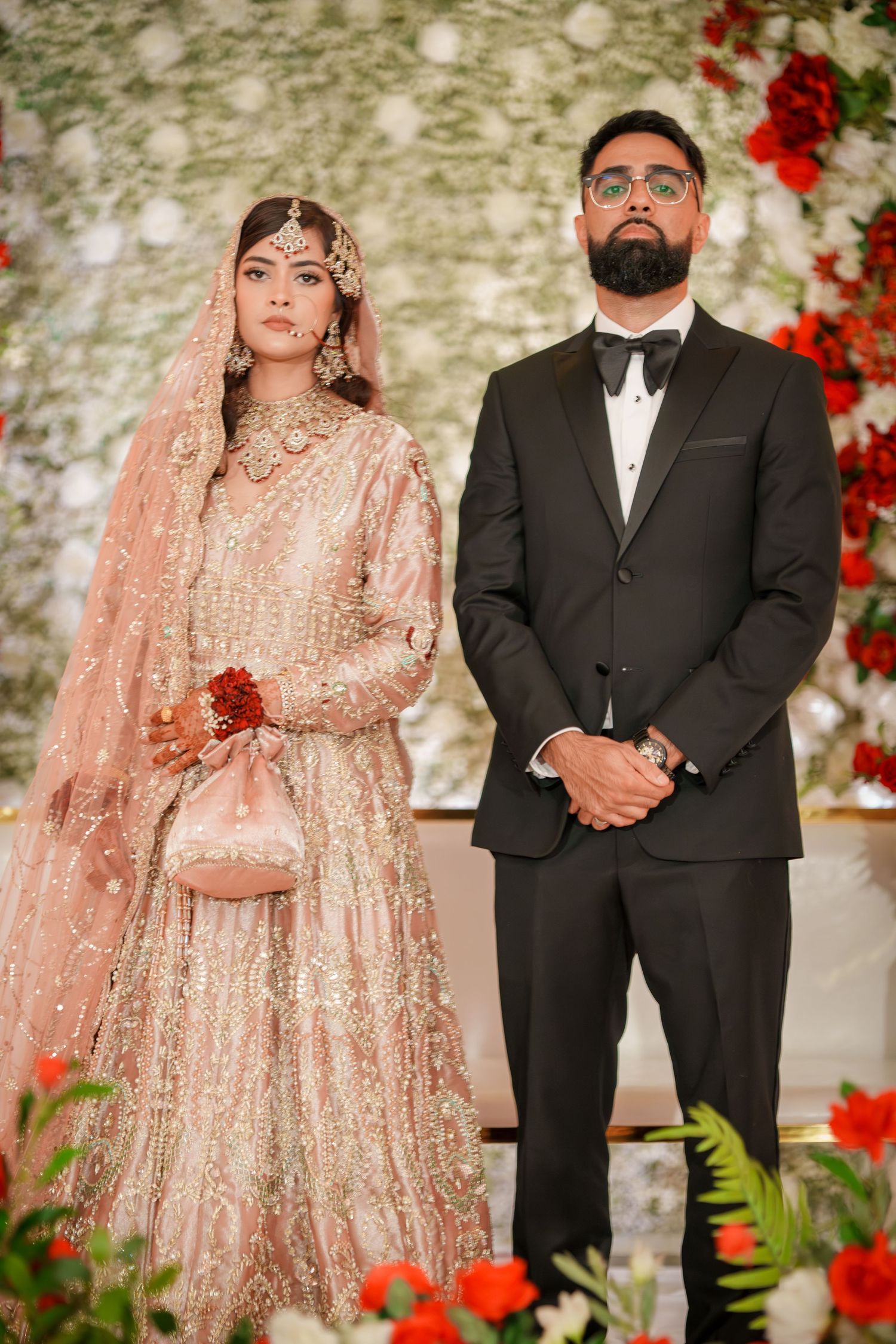 Wedding couple poses against floral backdrop with red roses and elegant decor in traditional attire.
