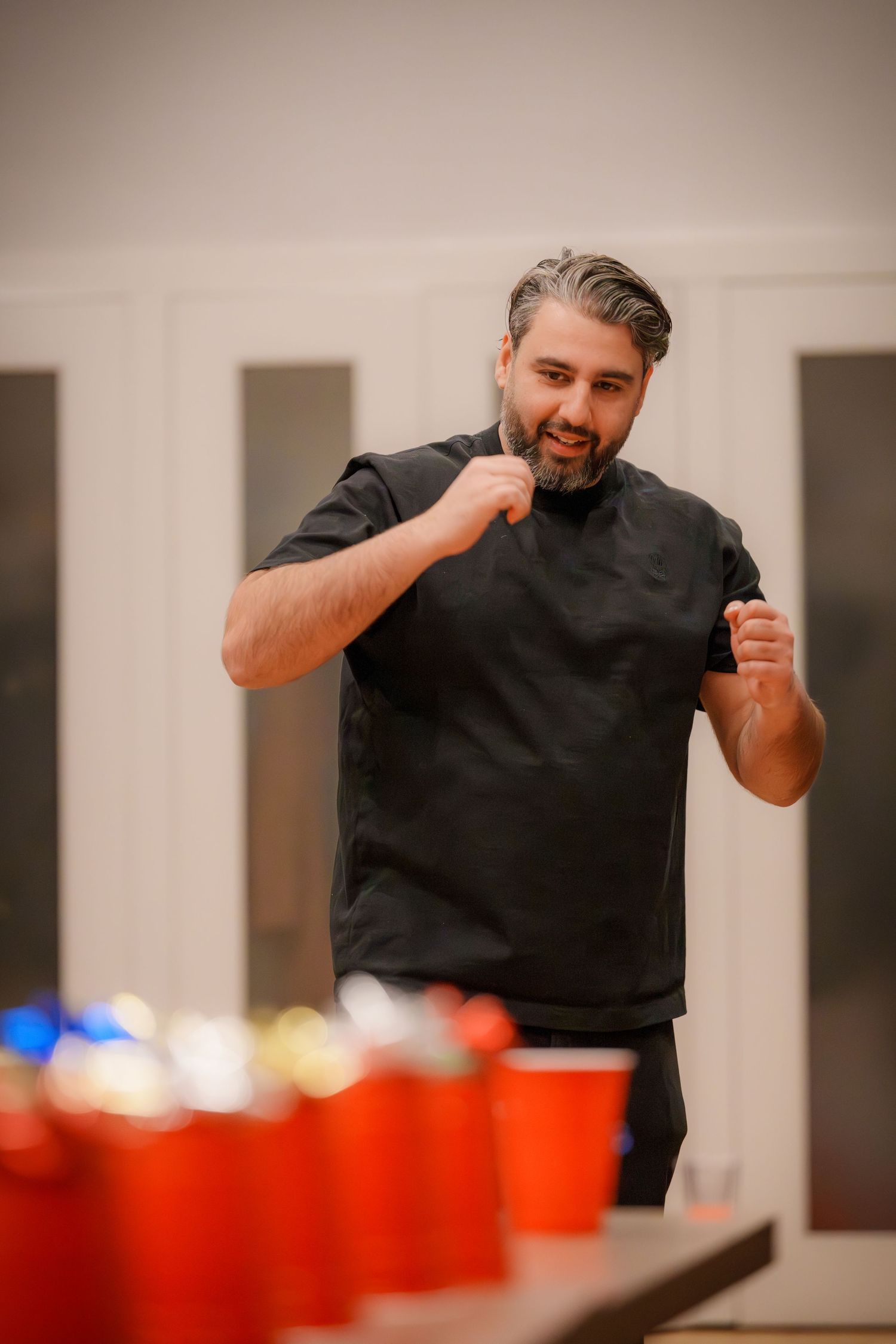 Person in black shirt playing beer pong game with red cups arranged on table.