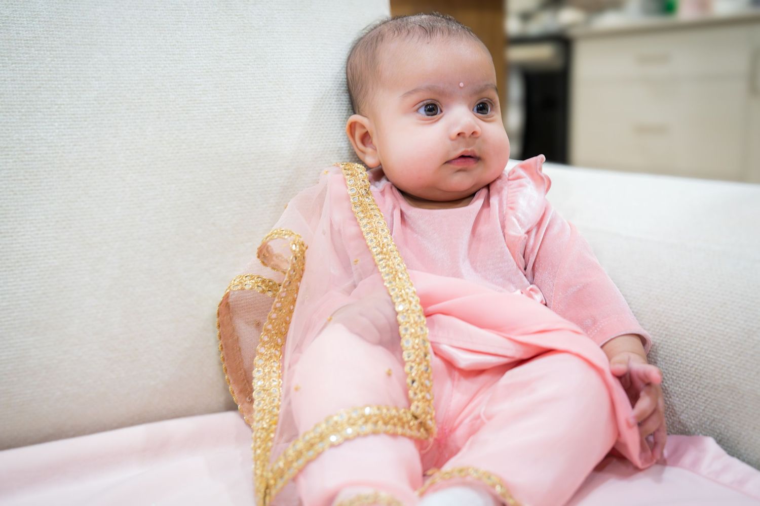 Adorable baby girl in pink traditional outfit sitting on white sofa, looking up curiously.
