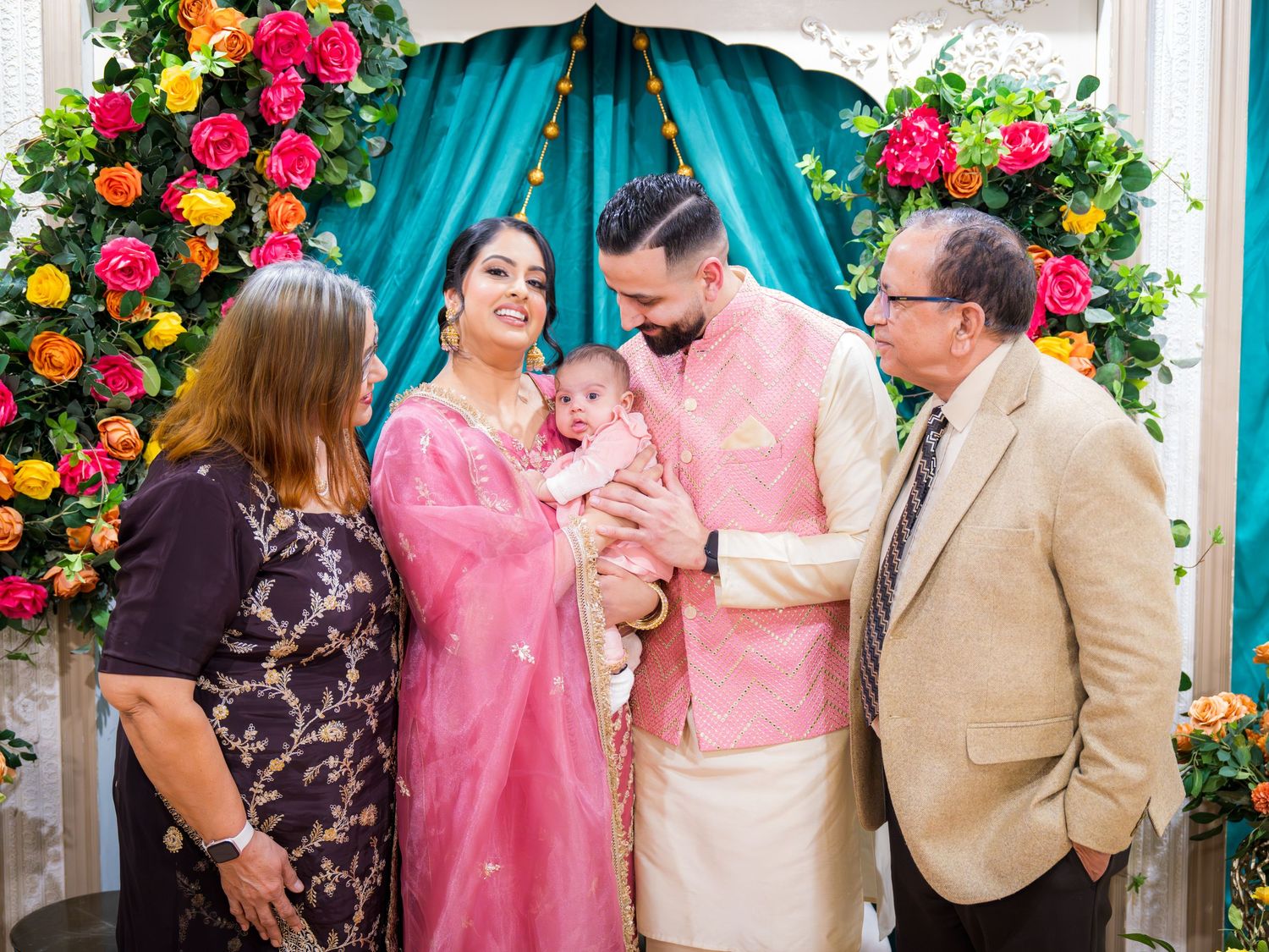 Indian couple in pink traditional attire introducing newborn baby to elderly relatives at floral decorated ceremony.