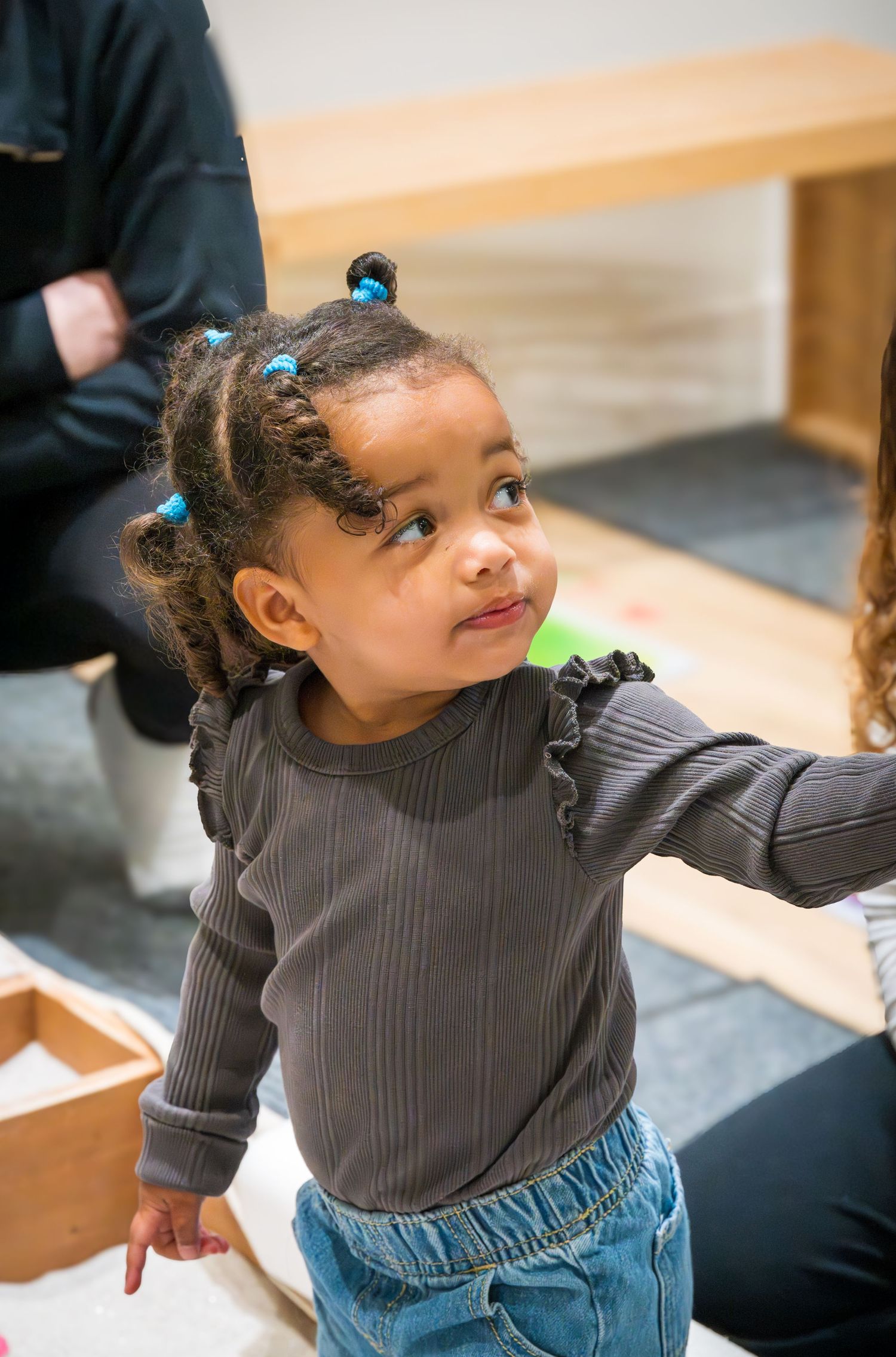 Cute young Black toddler girl with blue hair ties wearing a gray ruffled long-sleeve shirt and jeans indoors.