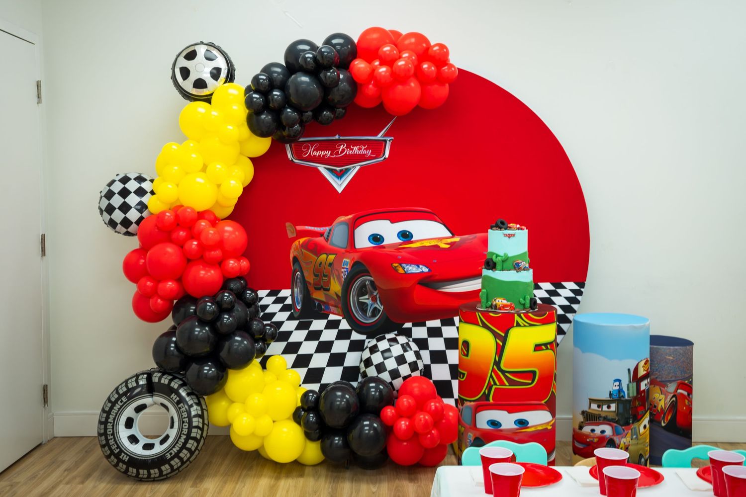 Cars themed birthday party decoration with Lightning McQueen backdrop, balloon garland in red, black, and yellow colors.