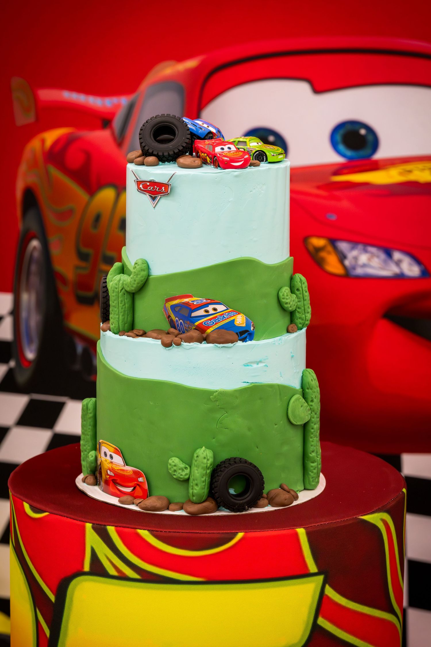 Two-tier Disney Cars themed birthday cake with Lightning McQueen decorations, cacti, and tires on a checkered table.